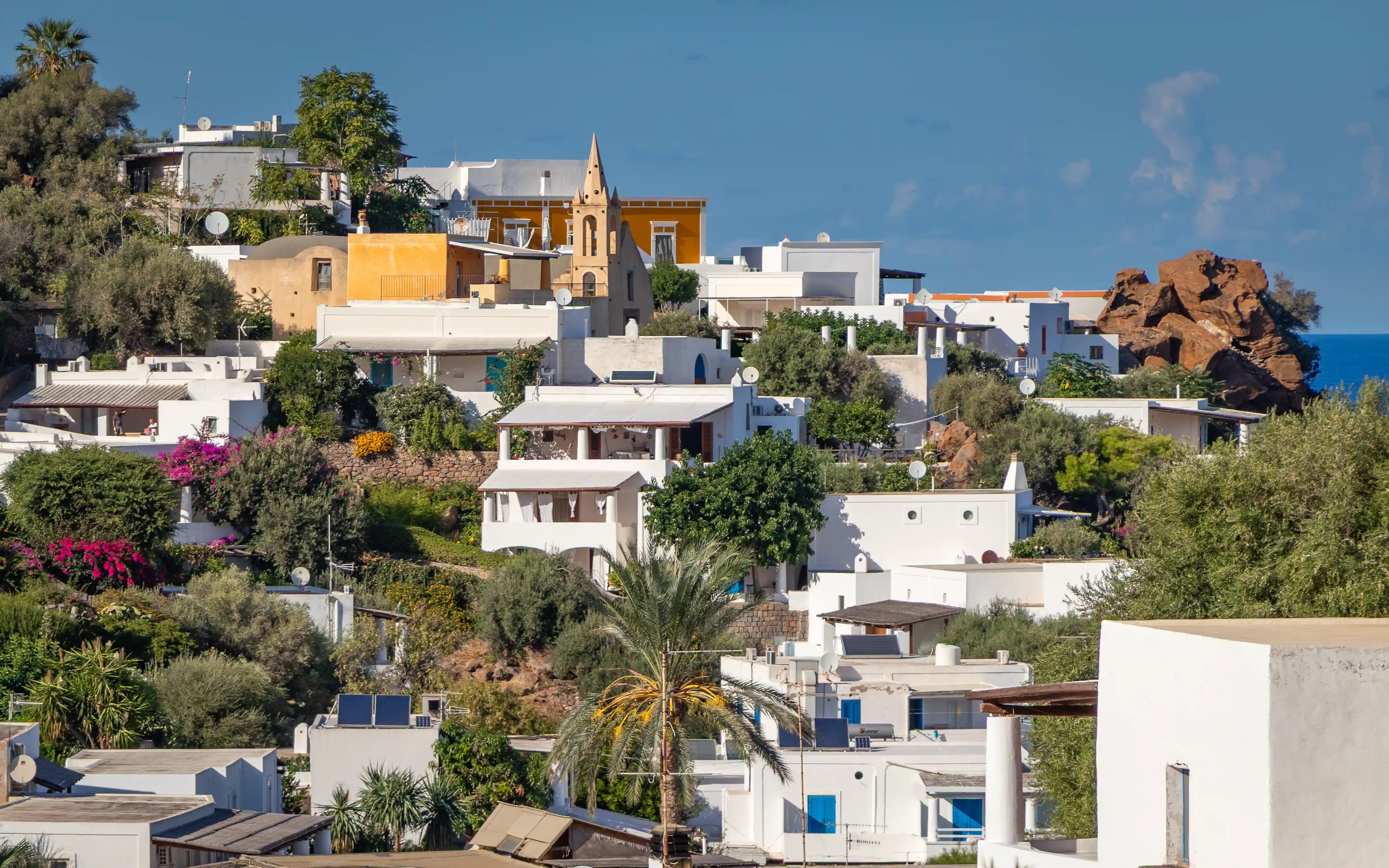 A hillside village with white buildings and a small chapel tower among trees and vegetation.