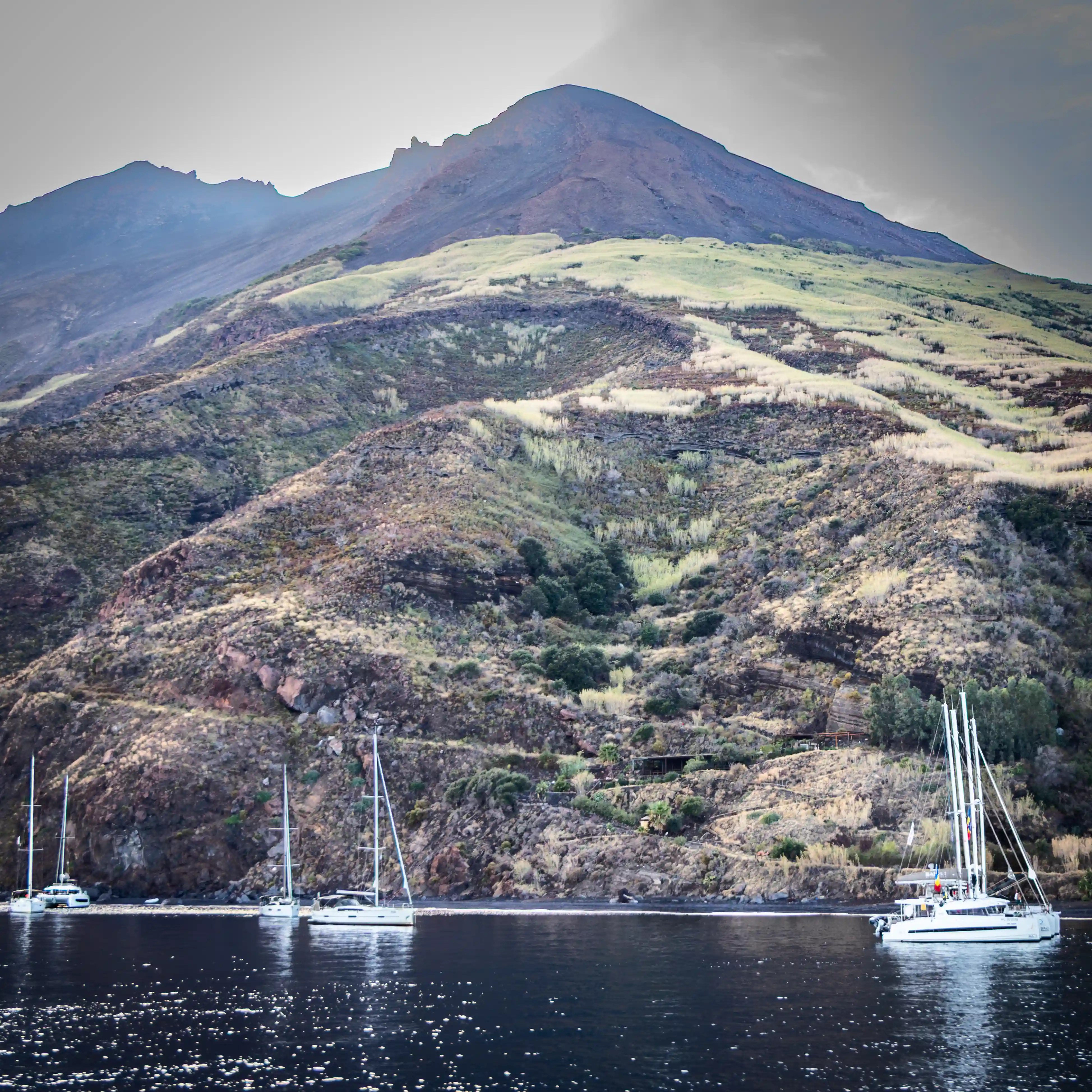 A volcanic hillside with sparse vegetation above calm water with several sailboats anchored nearby.