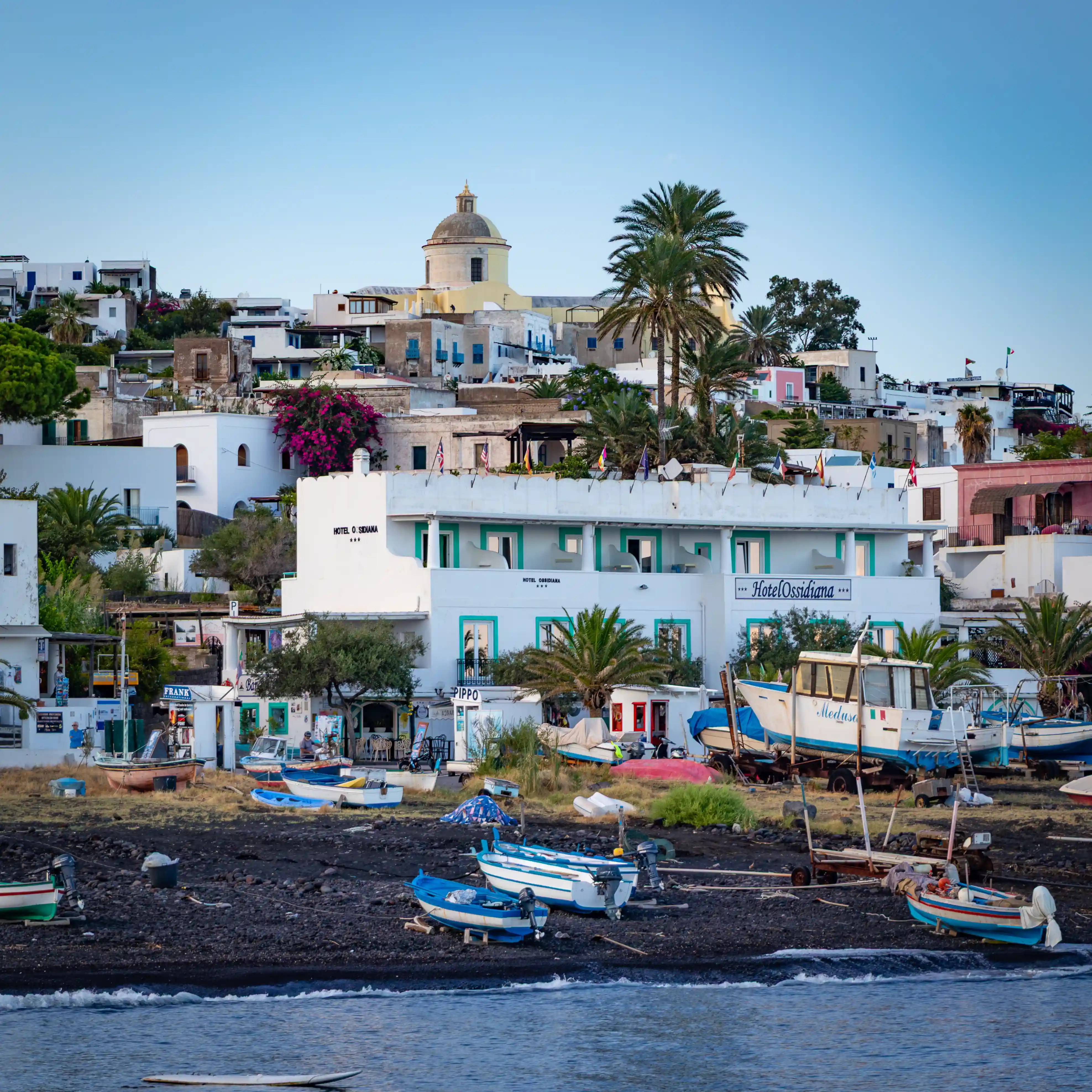 Small boats pulled onto a dark beach in front of white buildings and a hotel with palm trees behind.
