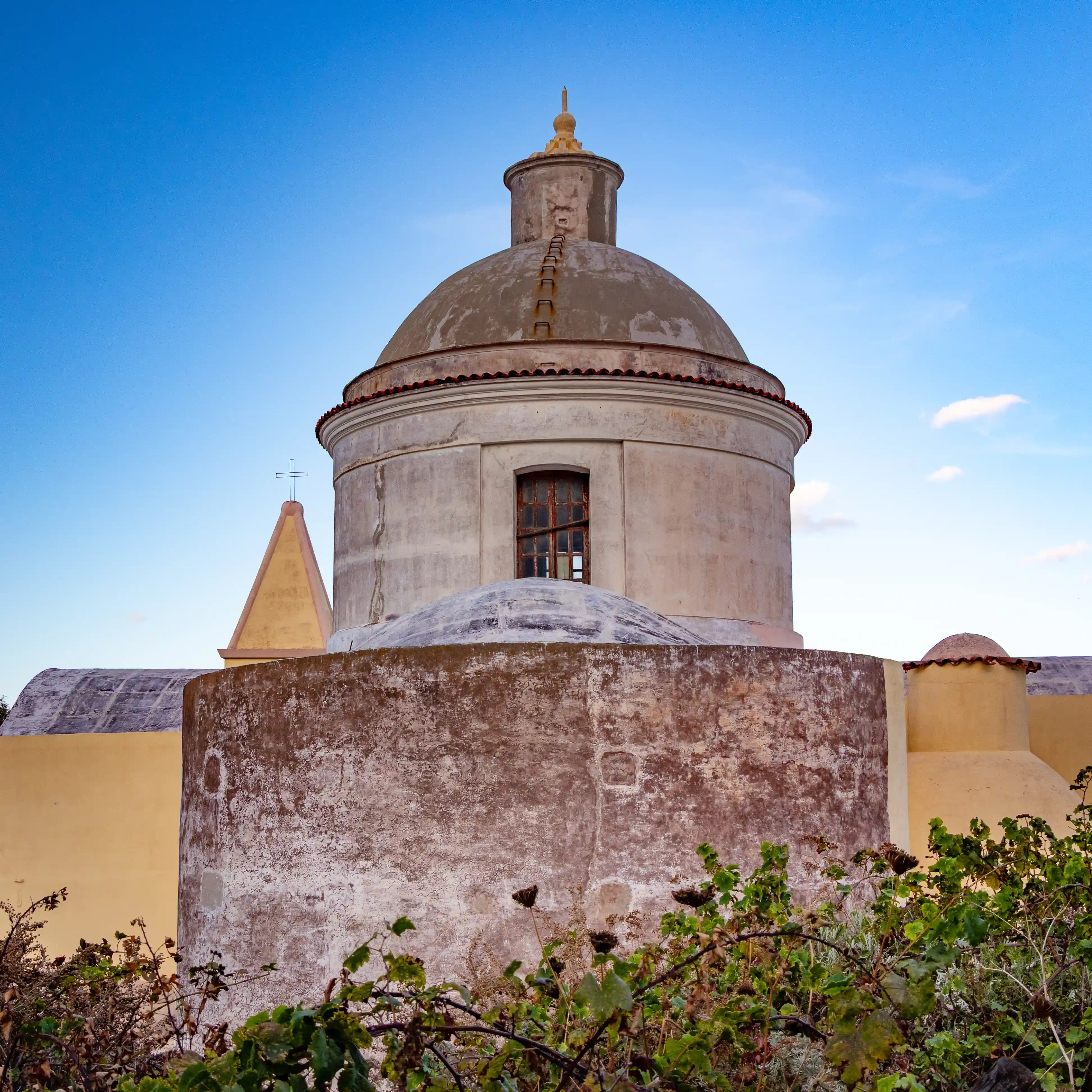 A round church dome with a small window and cross-topped structure behind it against a blue sky.