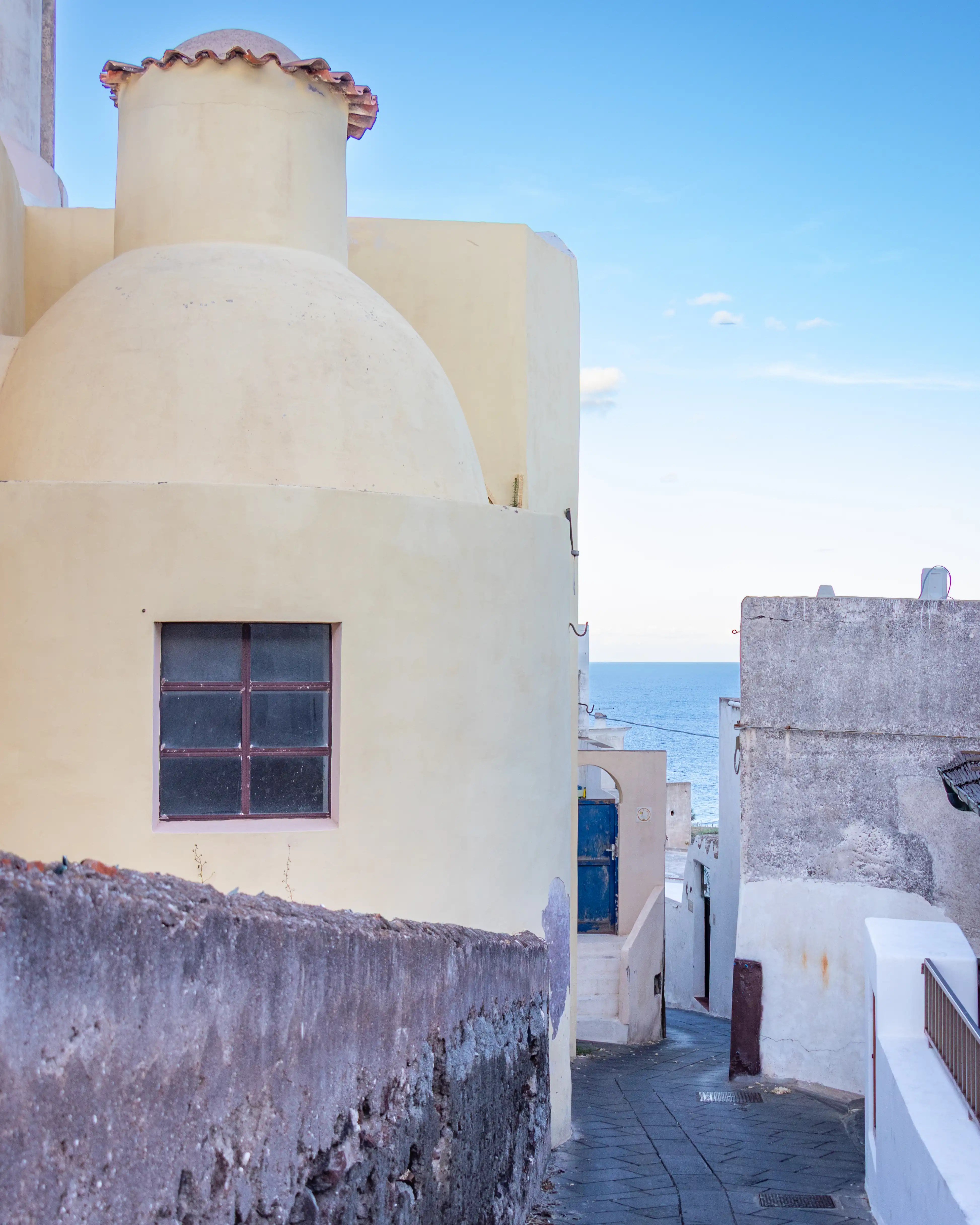 A narrow alley between light-colored buildings with a glimpse of the sea at the end.