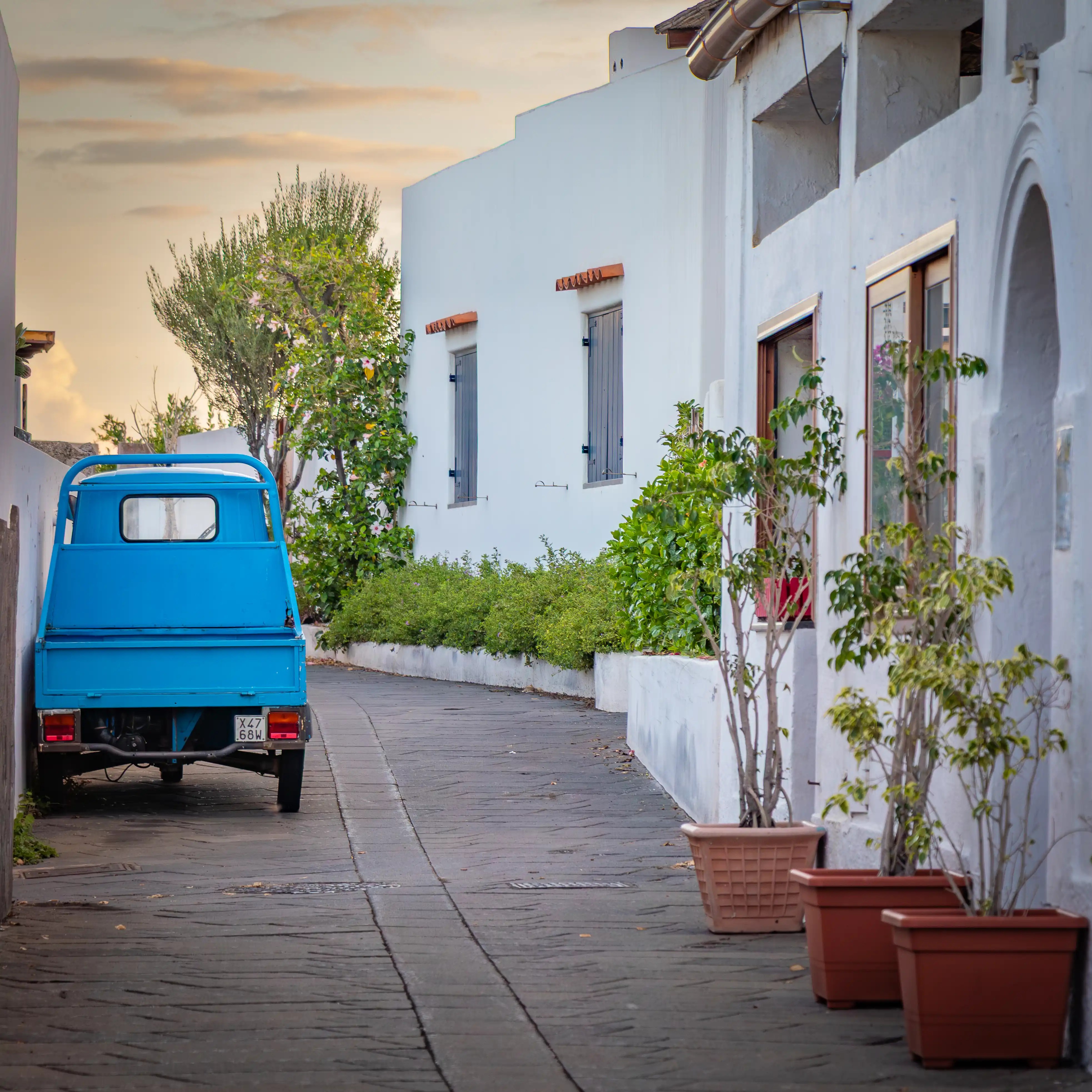 A small blue truck parked along a narrow street lined with white buildings and potted plants.