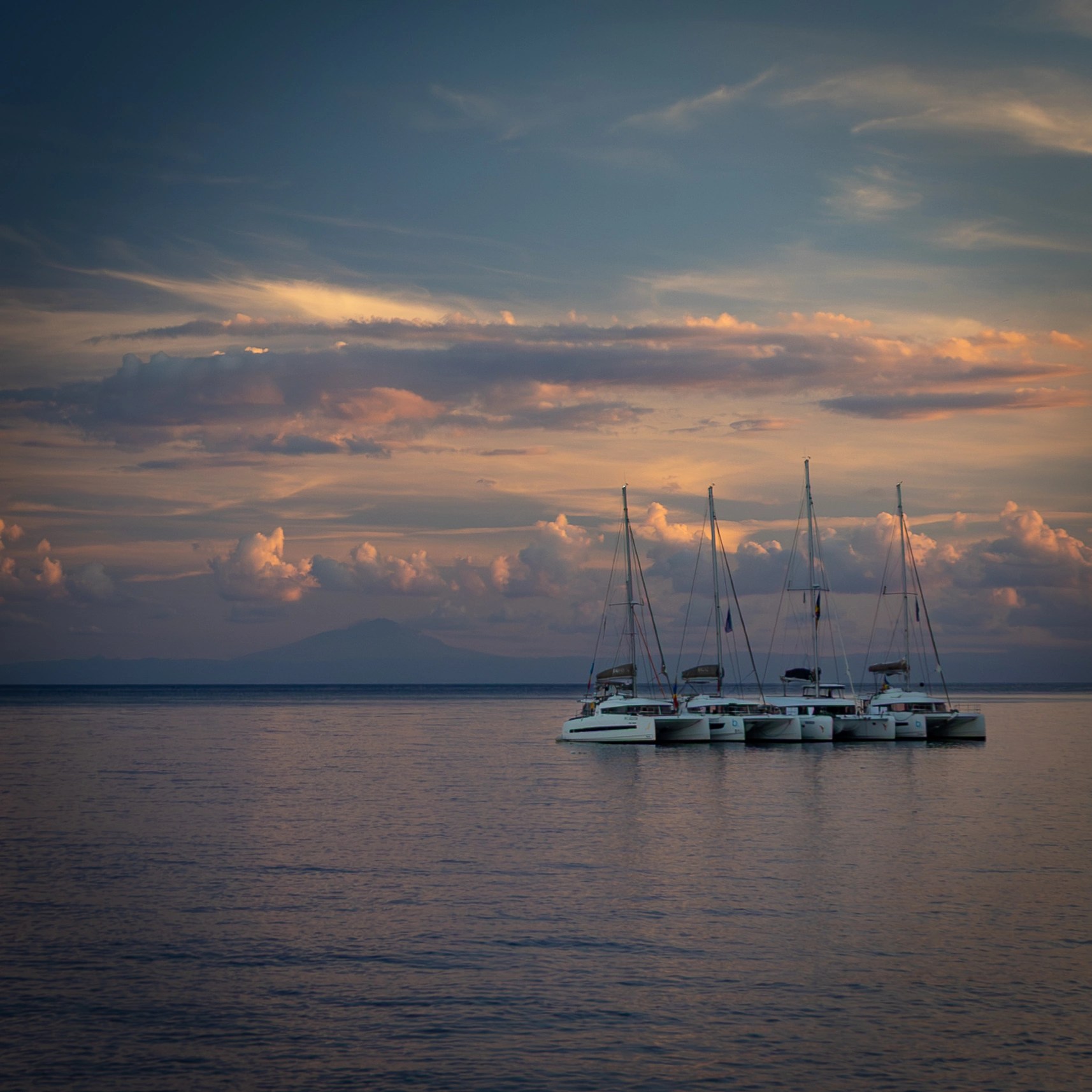Several sailboats anchored on calm water under a colorful sunset sky.