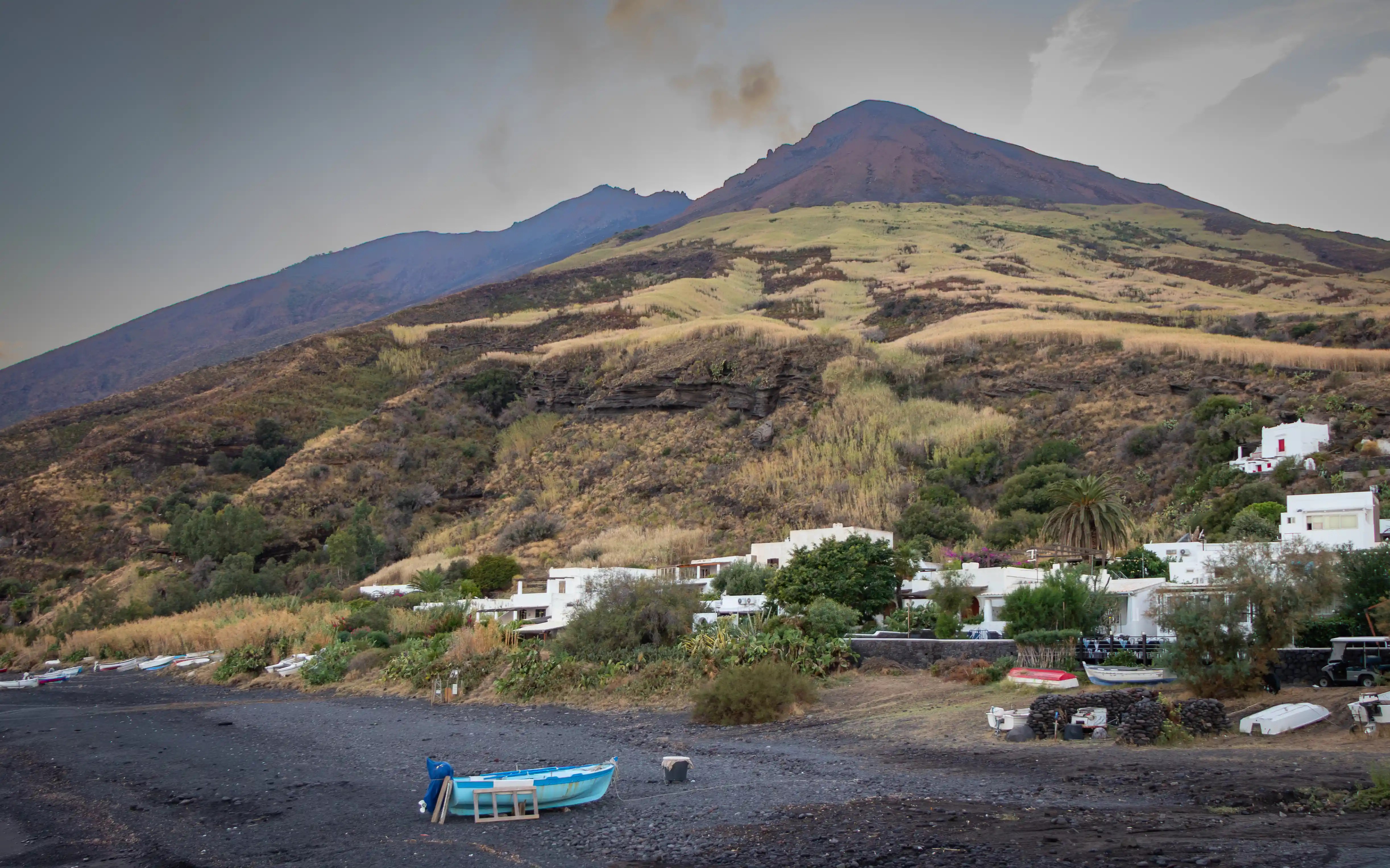 A dark shoreline with small boats and white buildings at the base of a volcanic mountain.