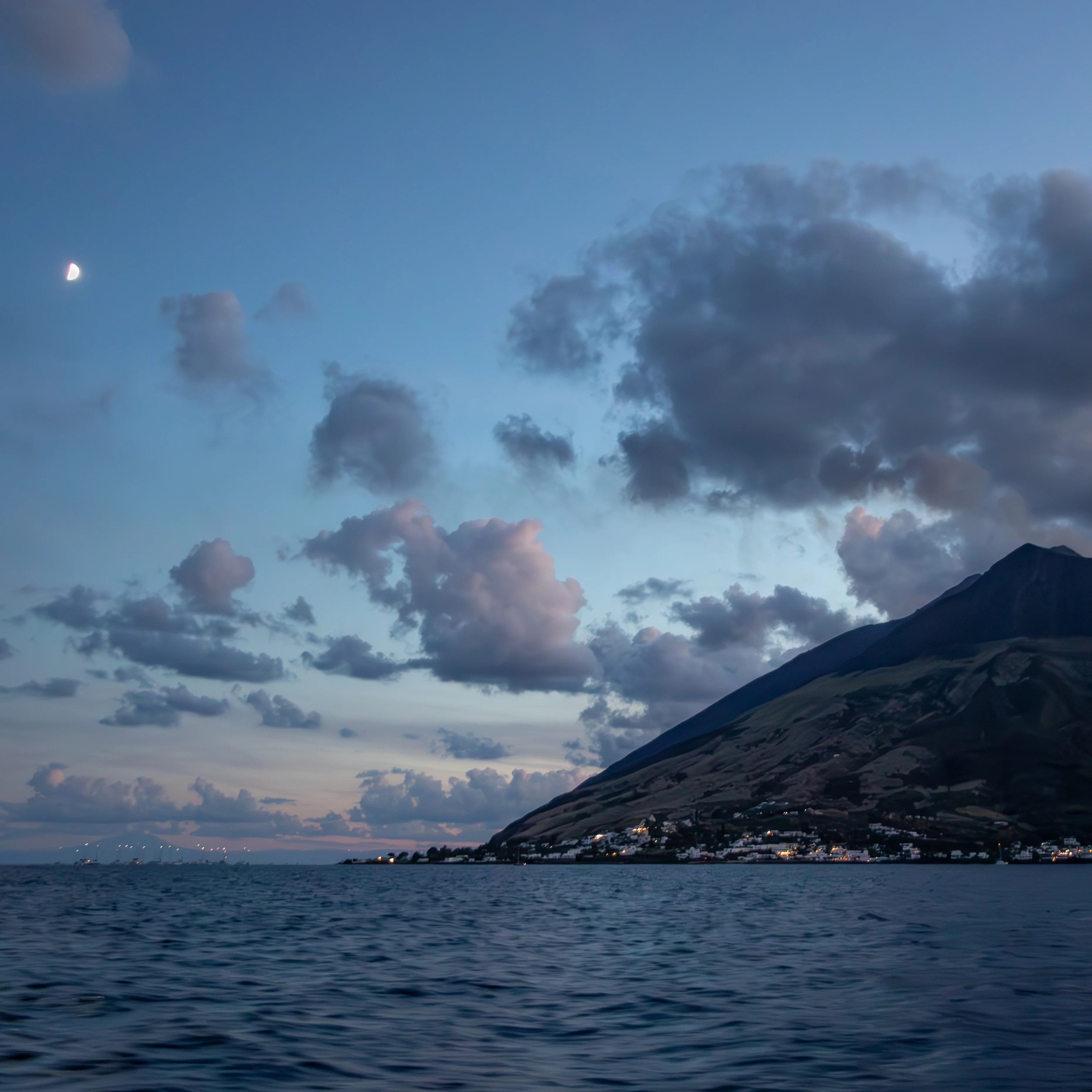 A coastline with buildings at dusk beneath a large mountain and a visible moon in the sky.