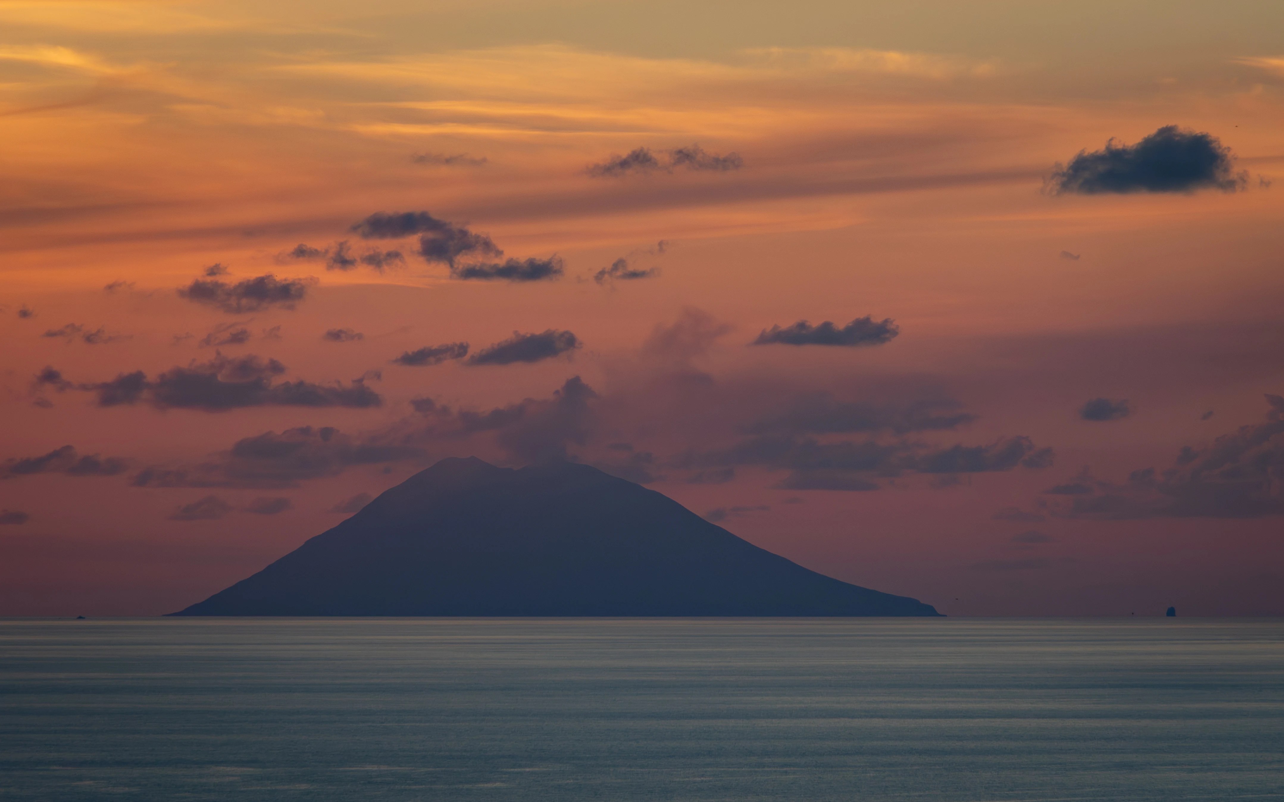 A dark silhouette of an island across calm water under a softly colored sunset sky.