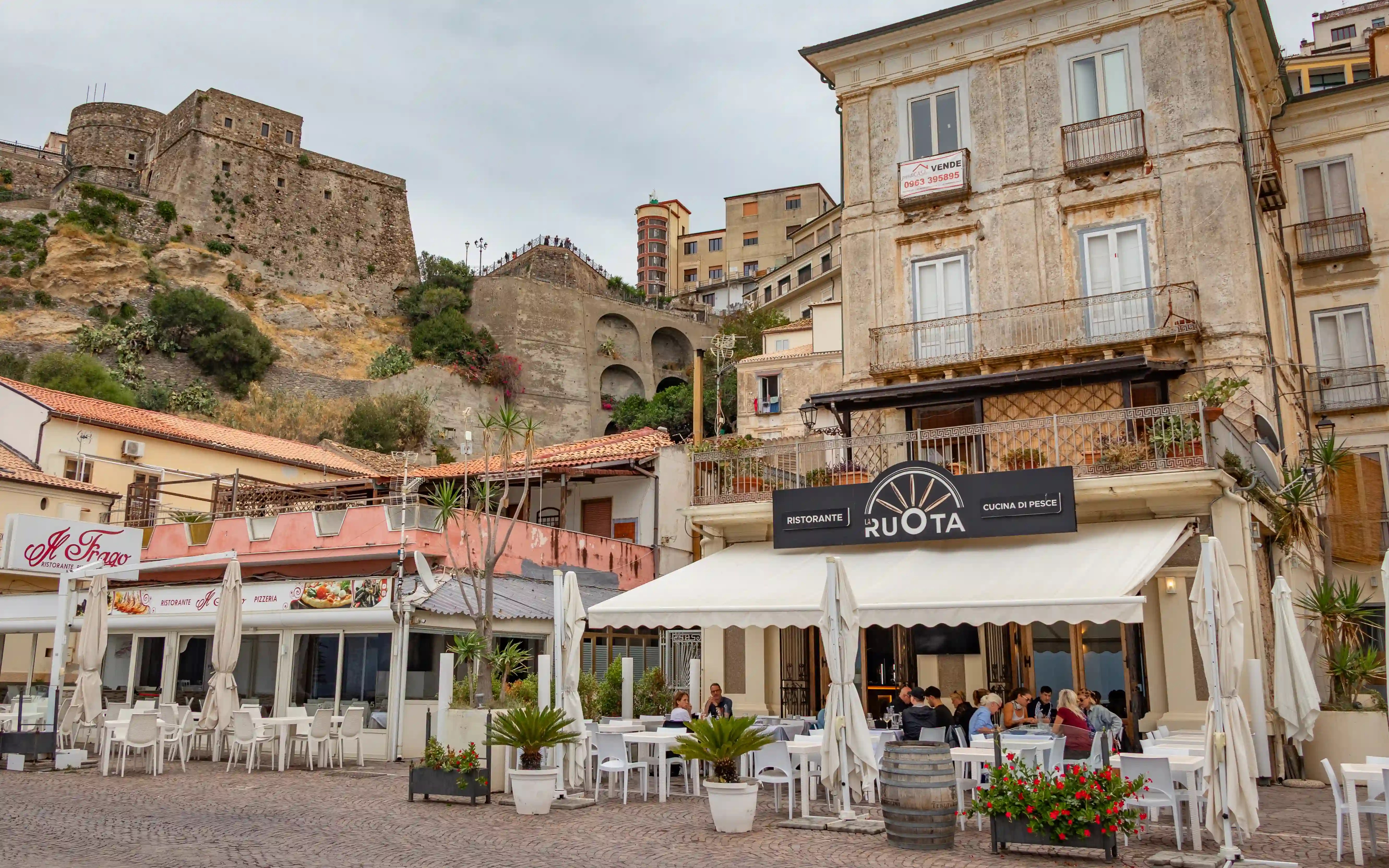 Restaurants with outdoor seating sit in front of older buildings, with a stone structure on a hill rising behind them.