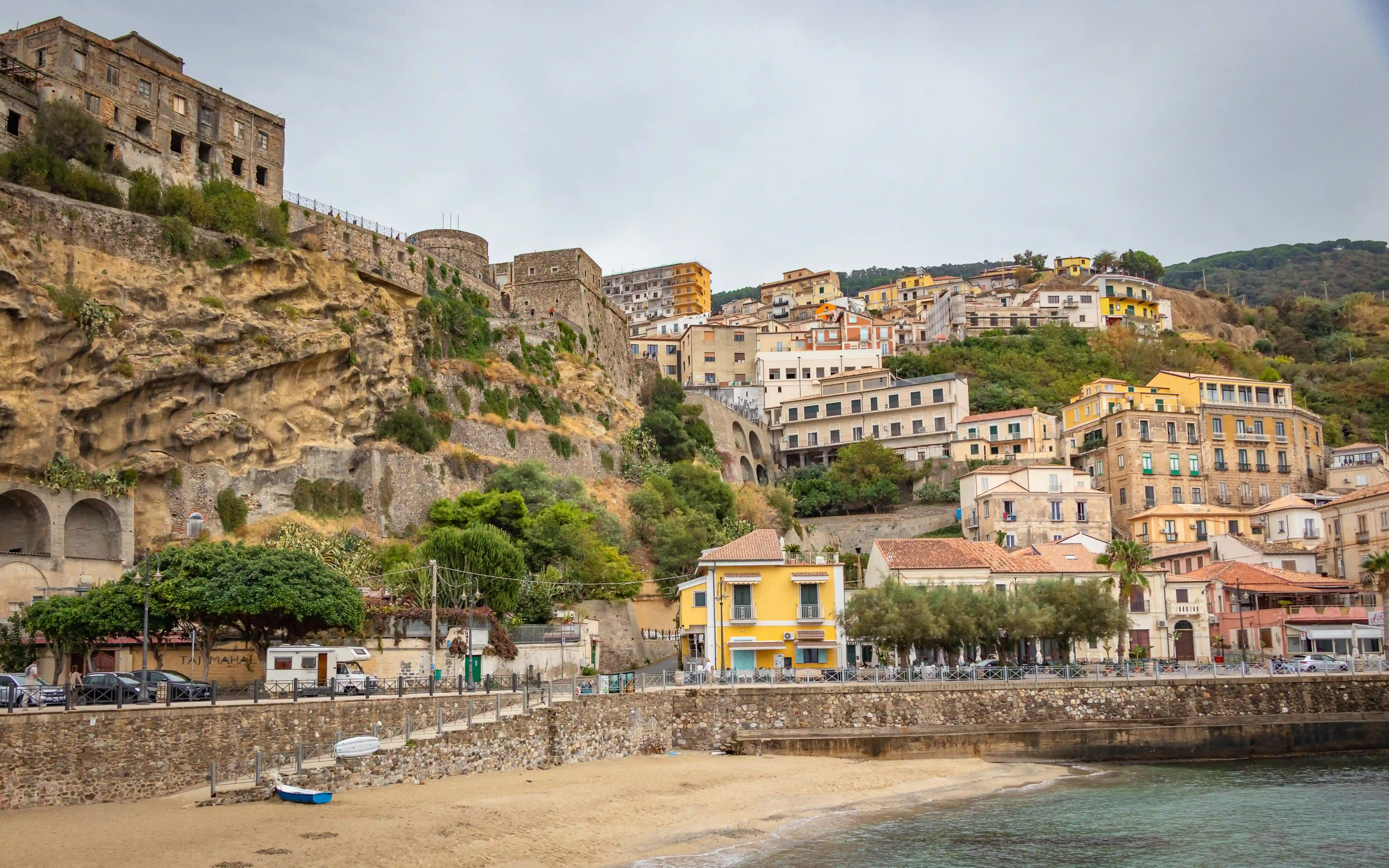 Coastal town built on a rocky cliff above a sandy beach with buildings and a stone wall along the shoreline.