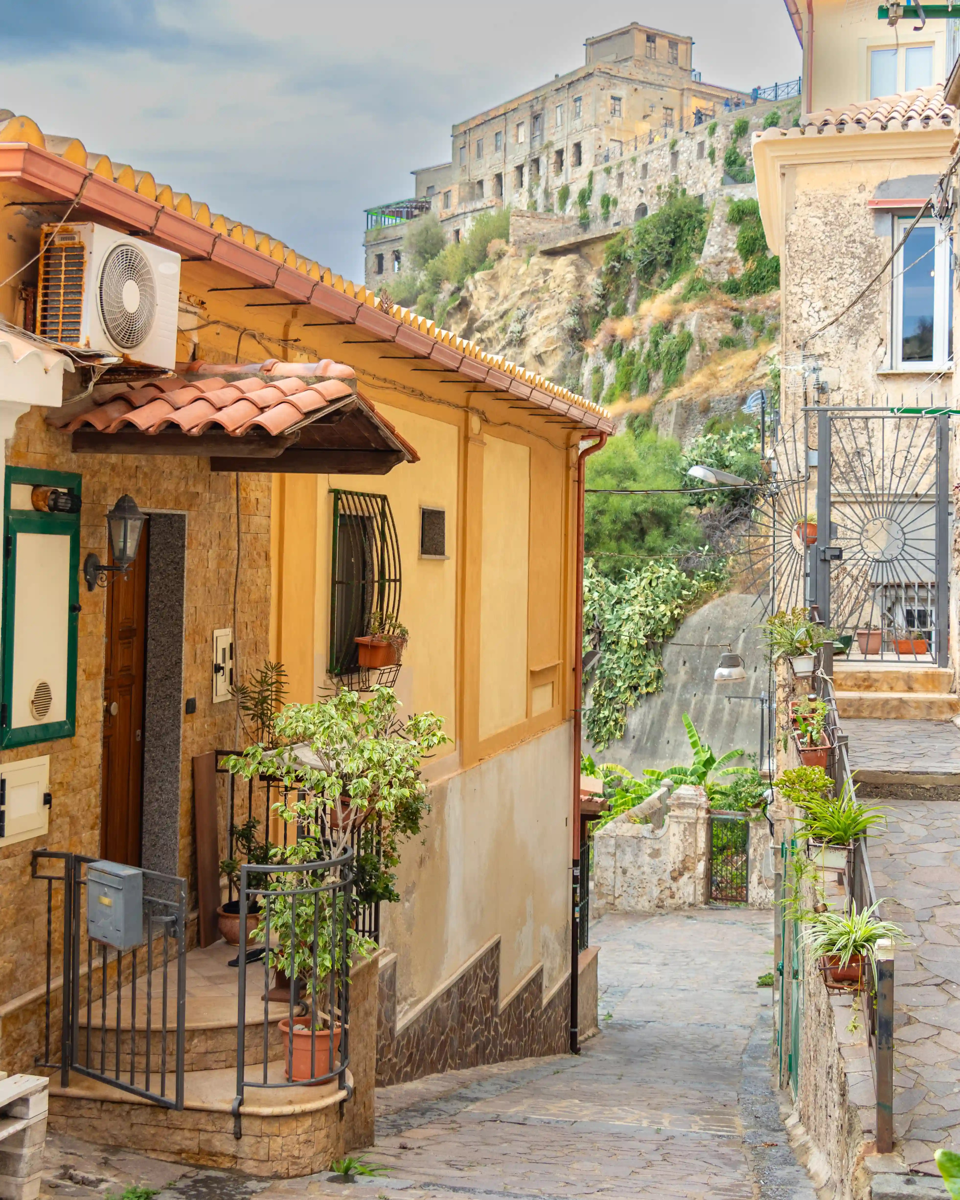 Narrow paved alley between houses with potted plants, leading toward a hillside with a large stone building above.
