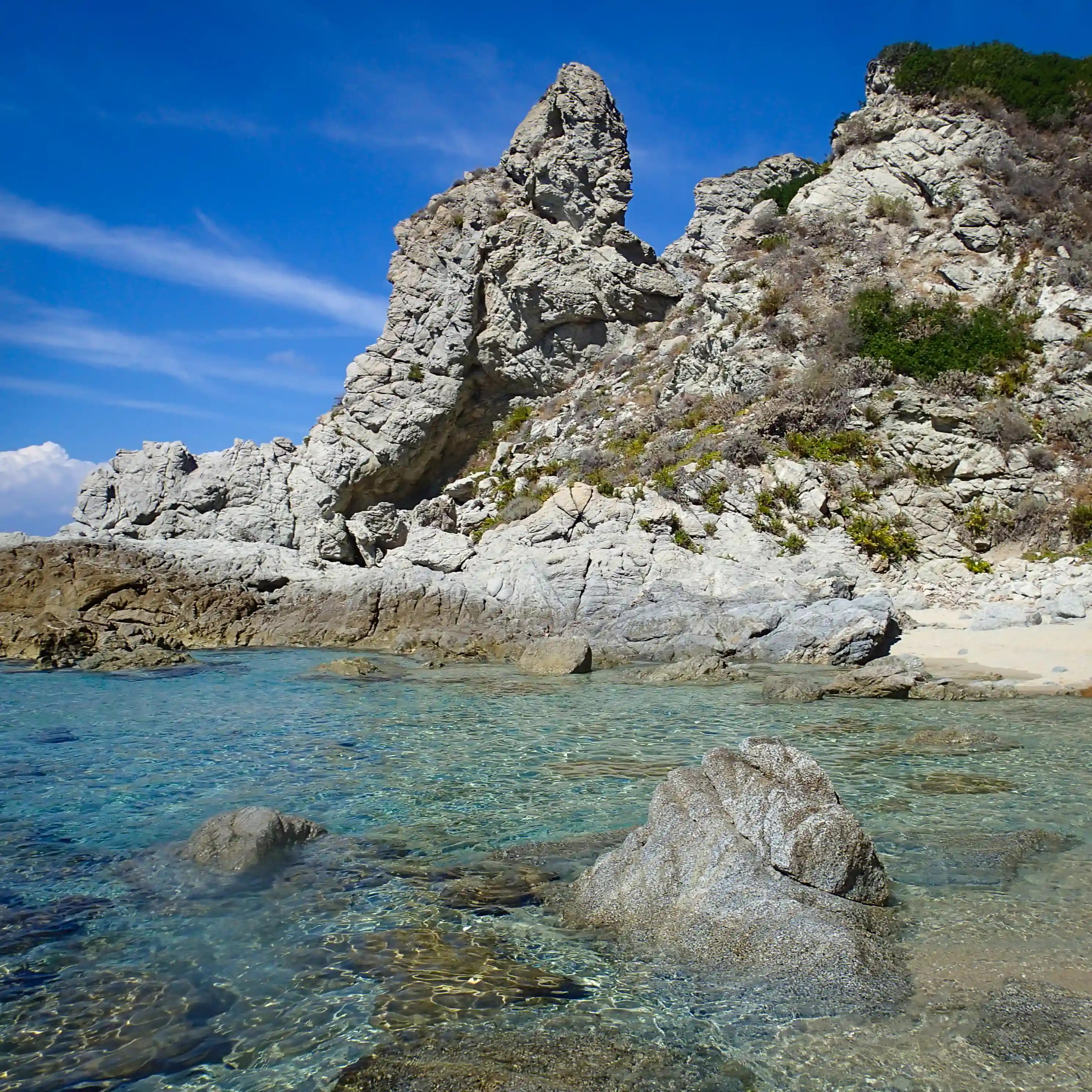 Clear shallow water with rocks in the foreground and jagged cliffs rising behind a small sandy cove.