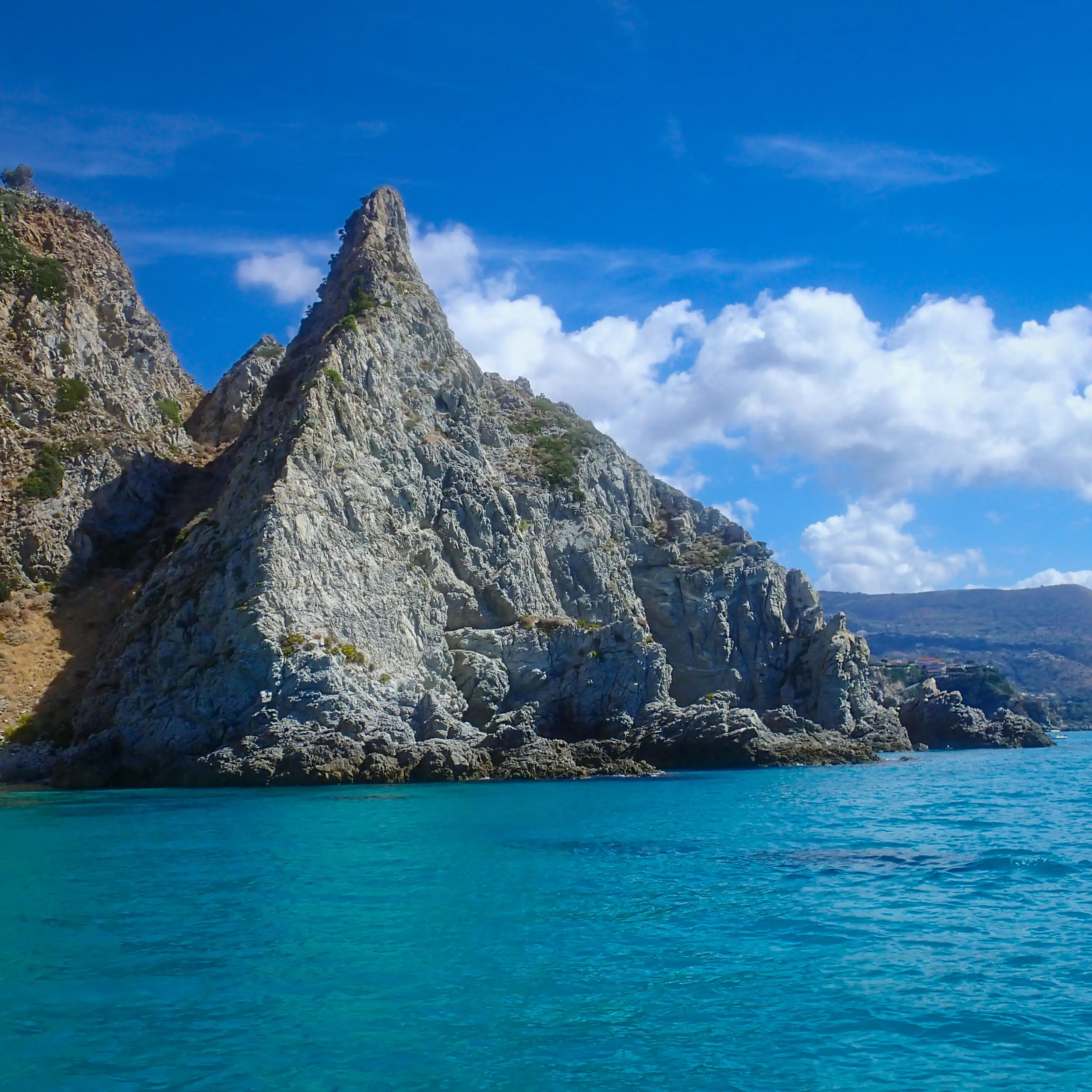 A tall, pointed rock formation rising from the sea with cliffs and clouds in the background.