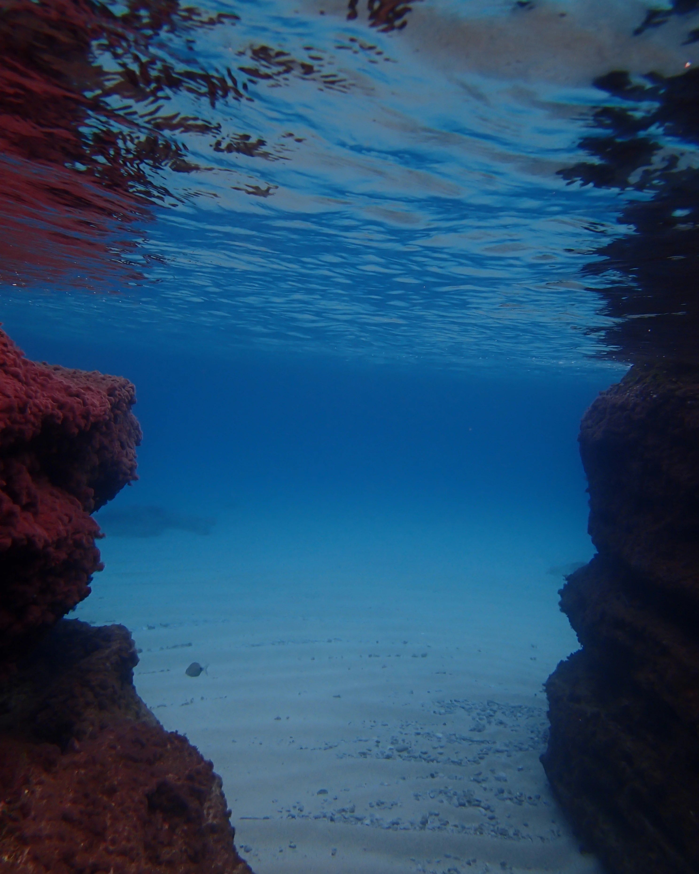 An underwater view between two rock formations showing a sandy seabed and light filtering through the water.