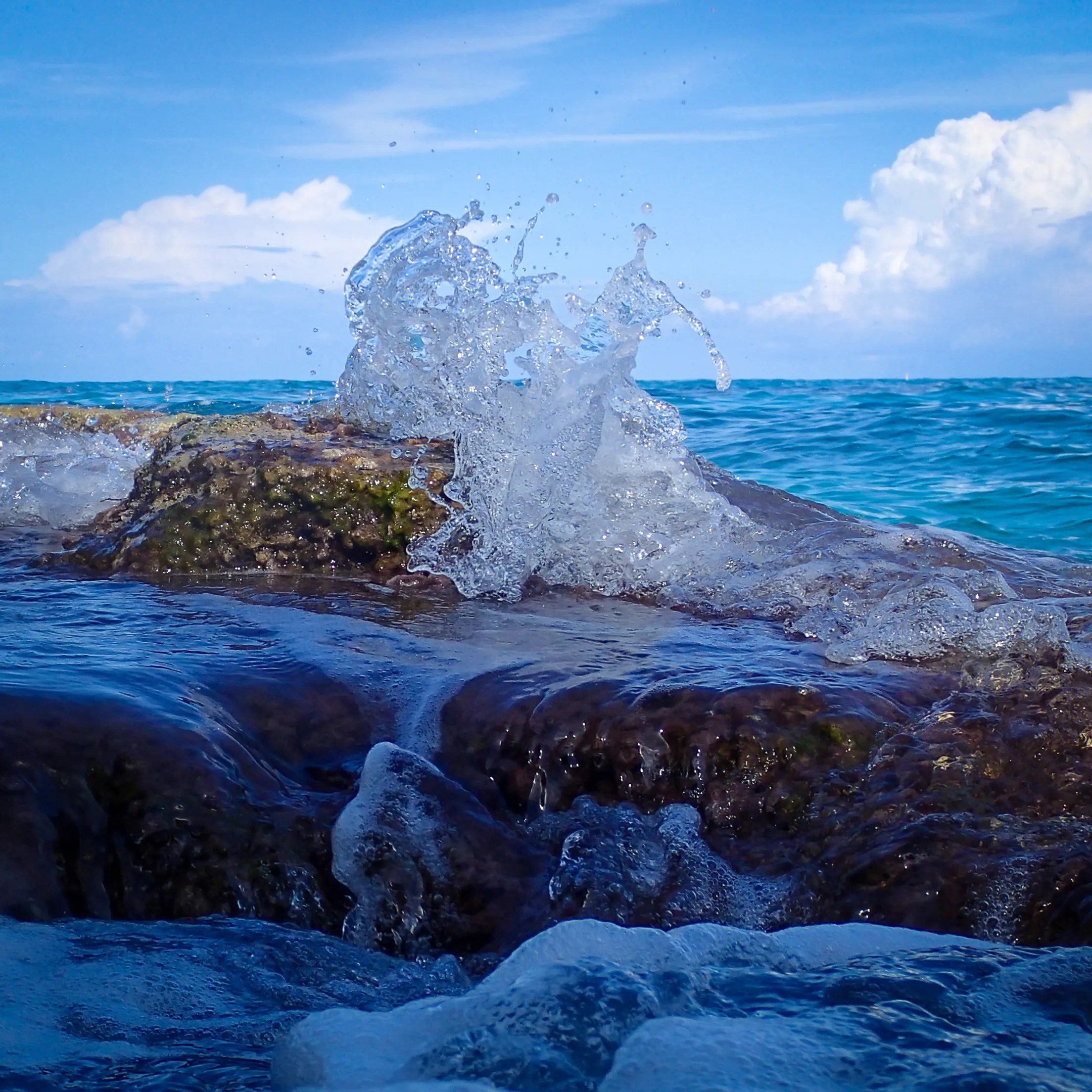 A splash of water breaking over rocks with the sea and sky in the background.