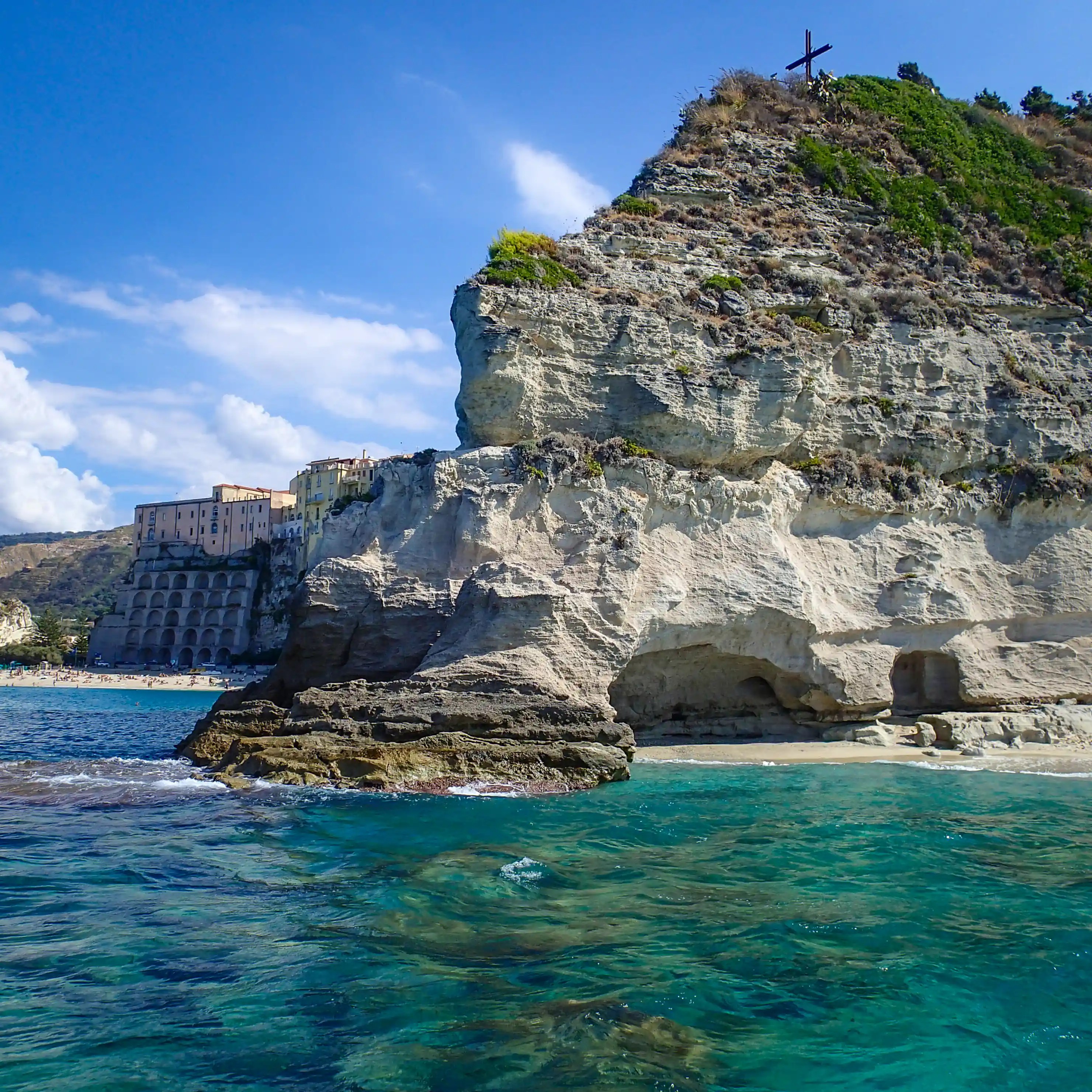 A rocky cliff at L’Isola in Tropea with a cross on top, turquoise water below, and buildings along the cliffs in the background.