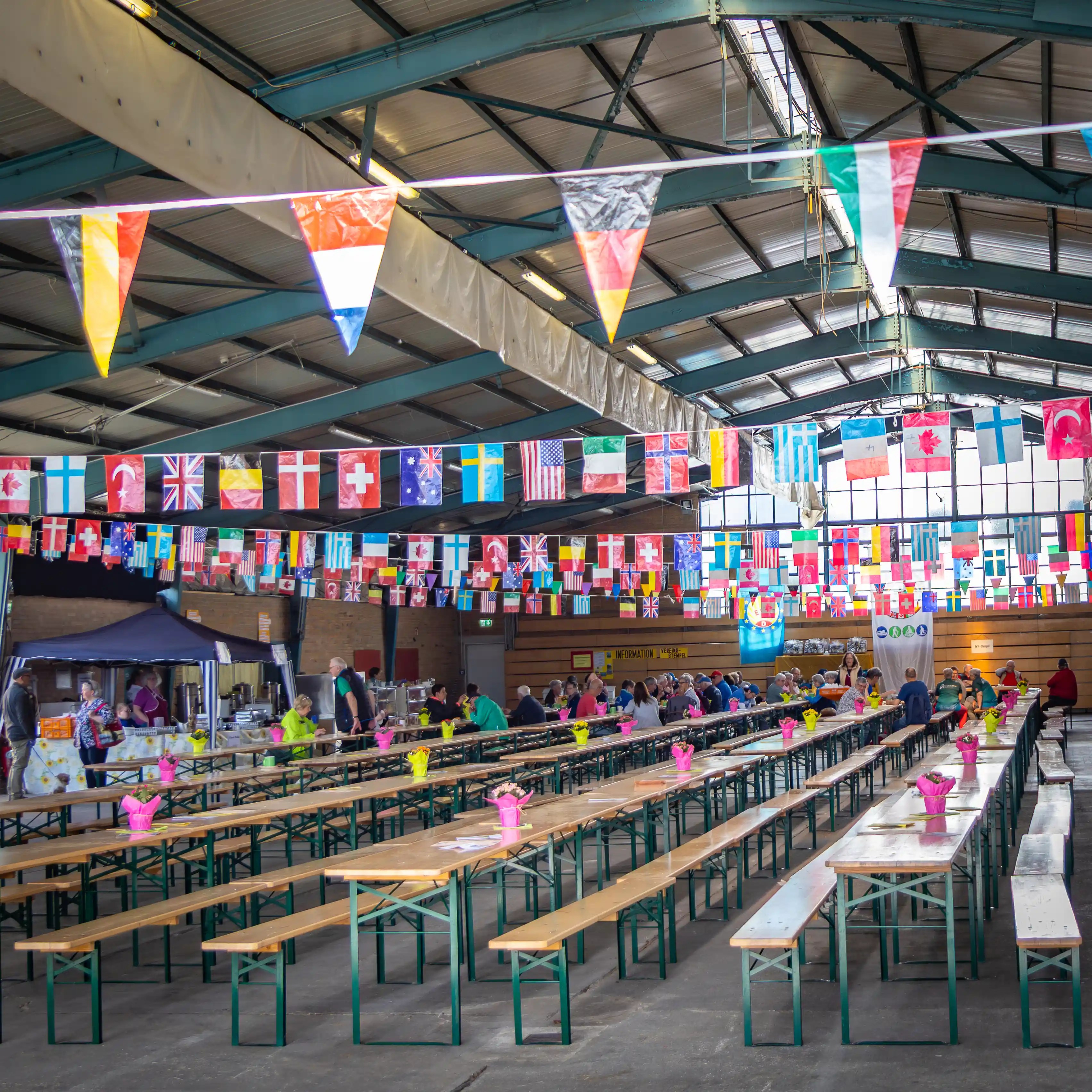 Long wooden tables sit beneath rows of international flags inside a large event hall.