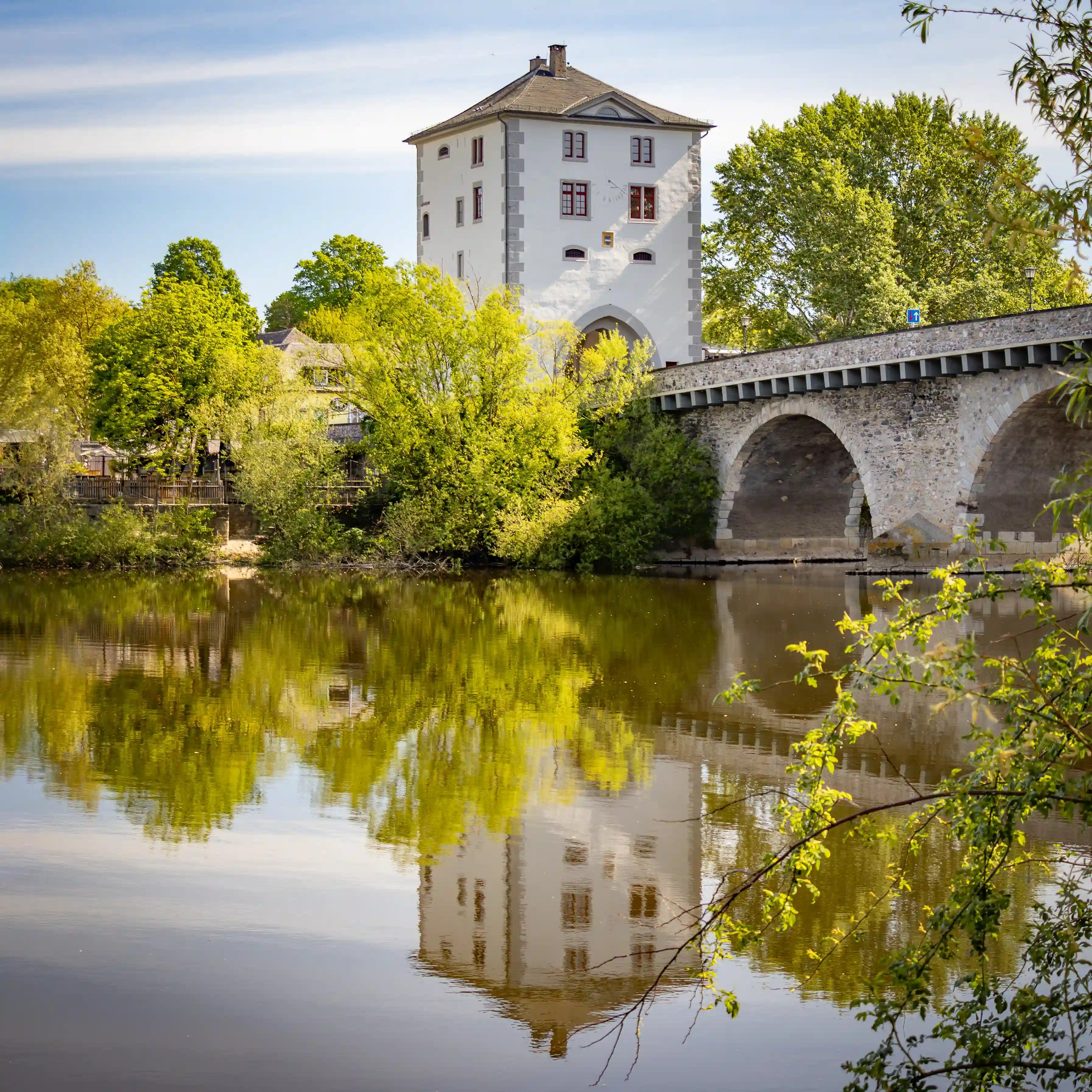 A white bridge tower and stone bridge reflect in the Lahn River surrounded by spring trees.