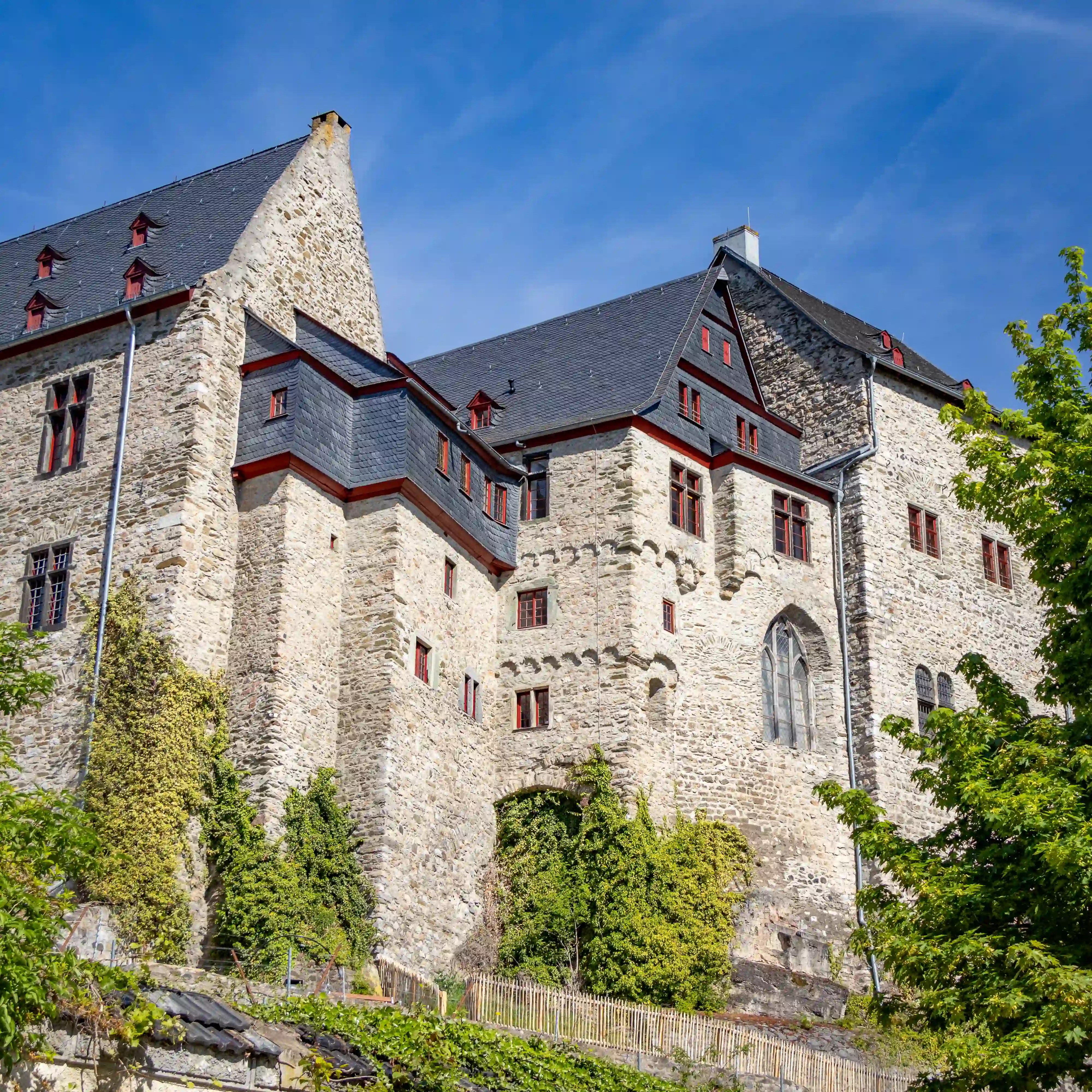 A large stone castle building with slate roofs and red-trimmed windows stands above greenery.