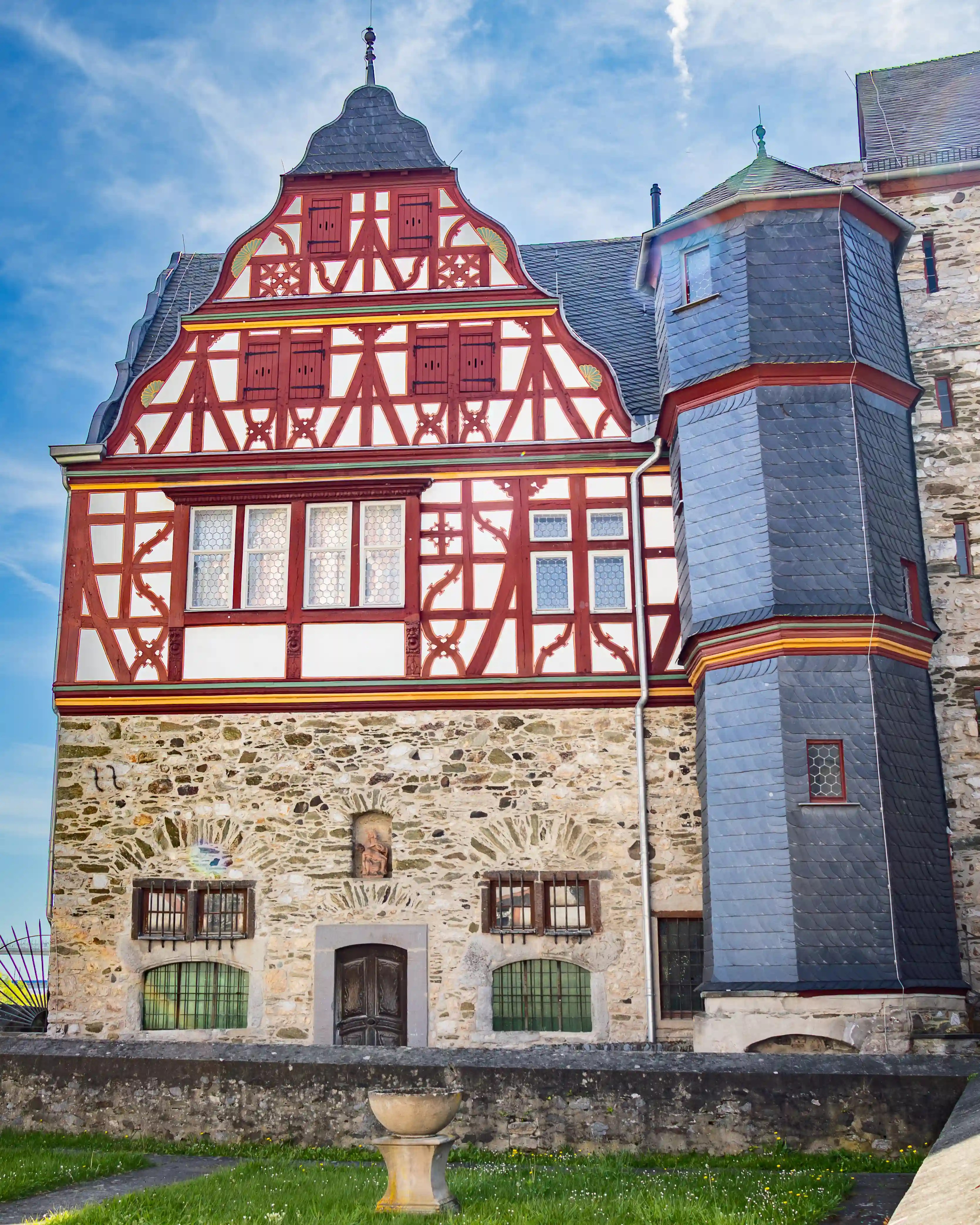 A red-and-white half-timbered building stands beside a slate-covered tower and stone wall.