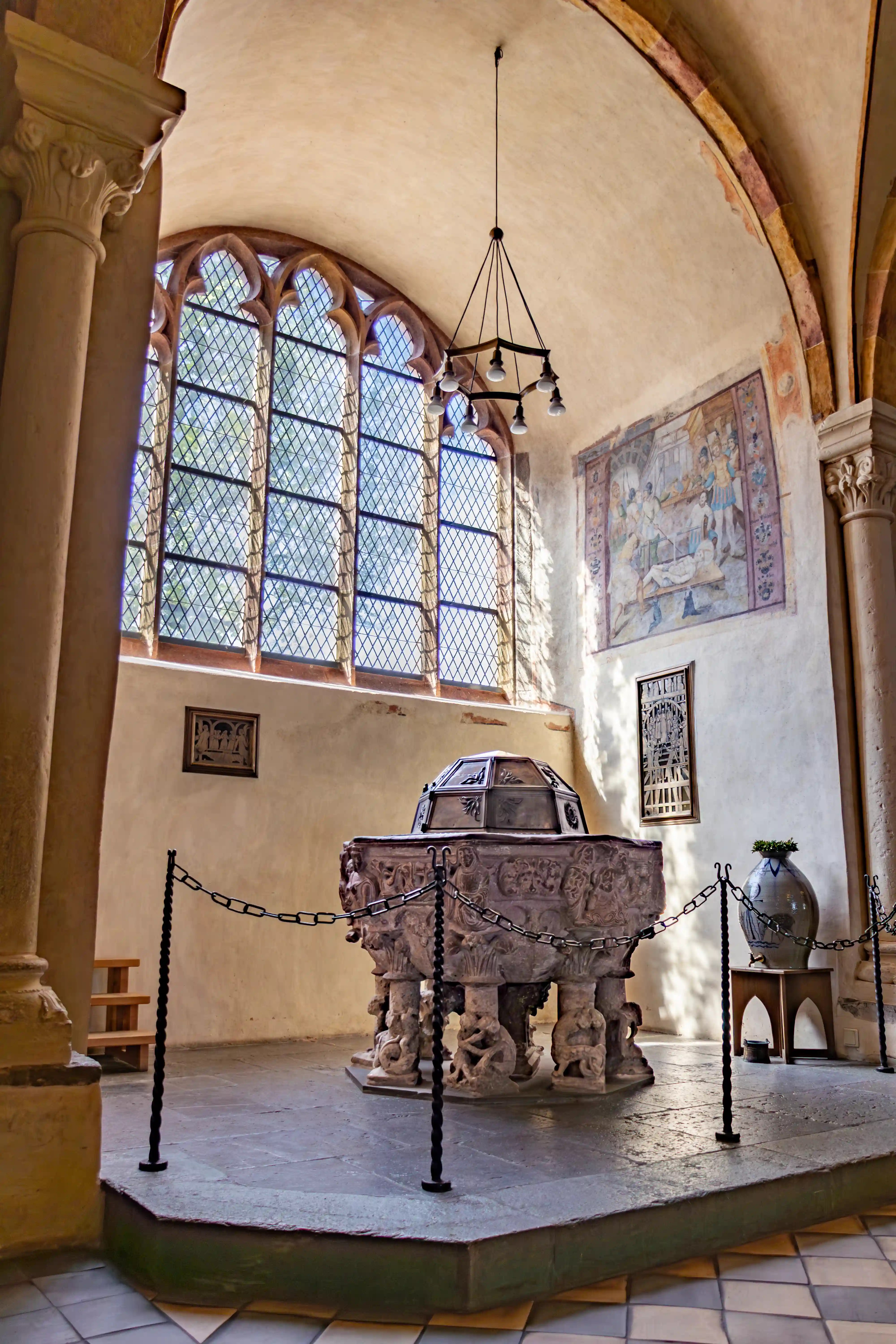 A carved baptismal font stands below tall windows and a wall painting inside a church.