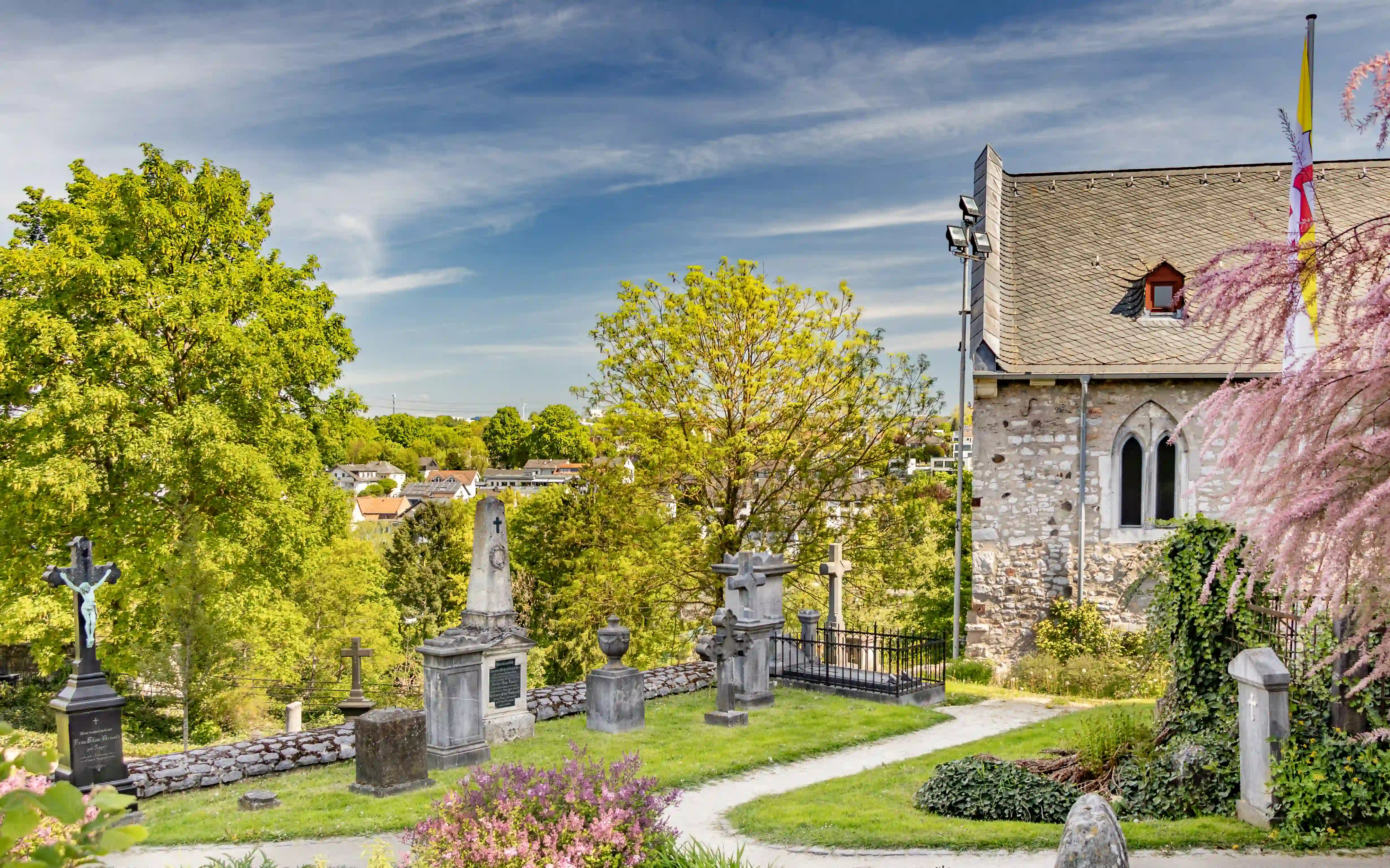 Gravestones and a small stone church building sit on a hillside with trees and town views.