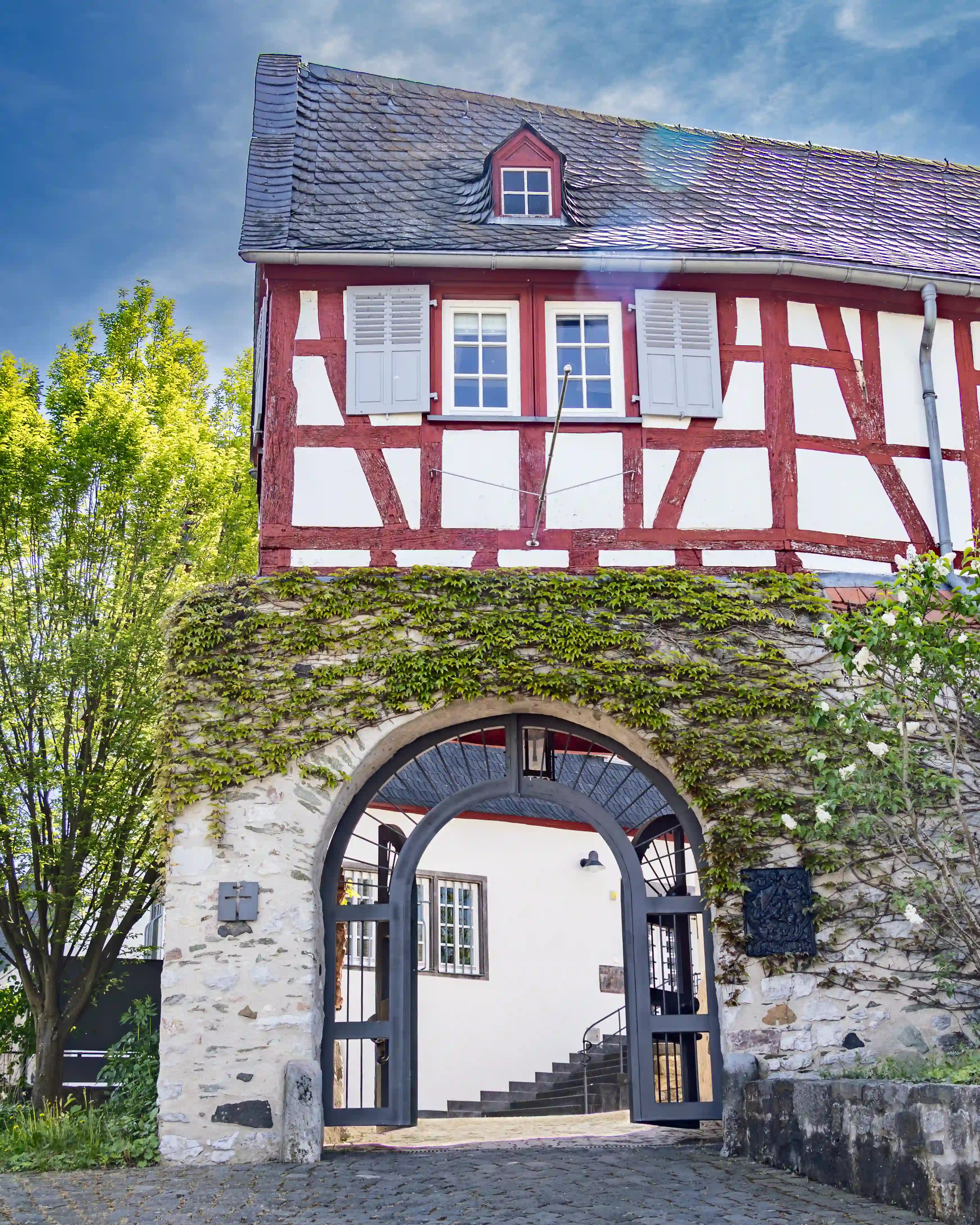 A red-and-white half-timbered building stands above an arched stone gateway covered with greenery.
