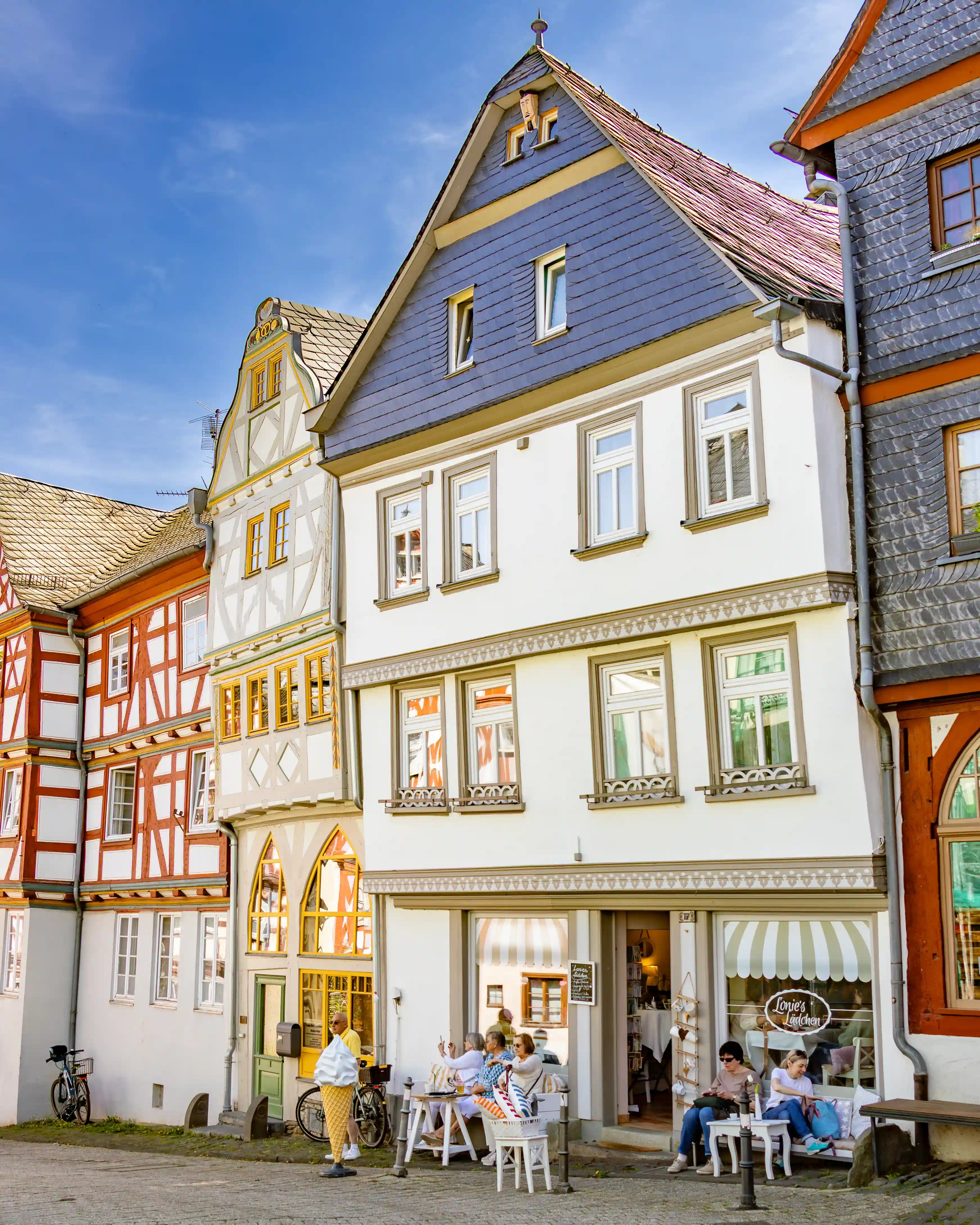 Several colorful buildings line a sunny street with people sitting outside a small shop.