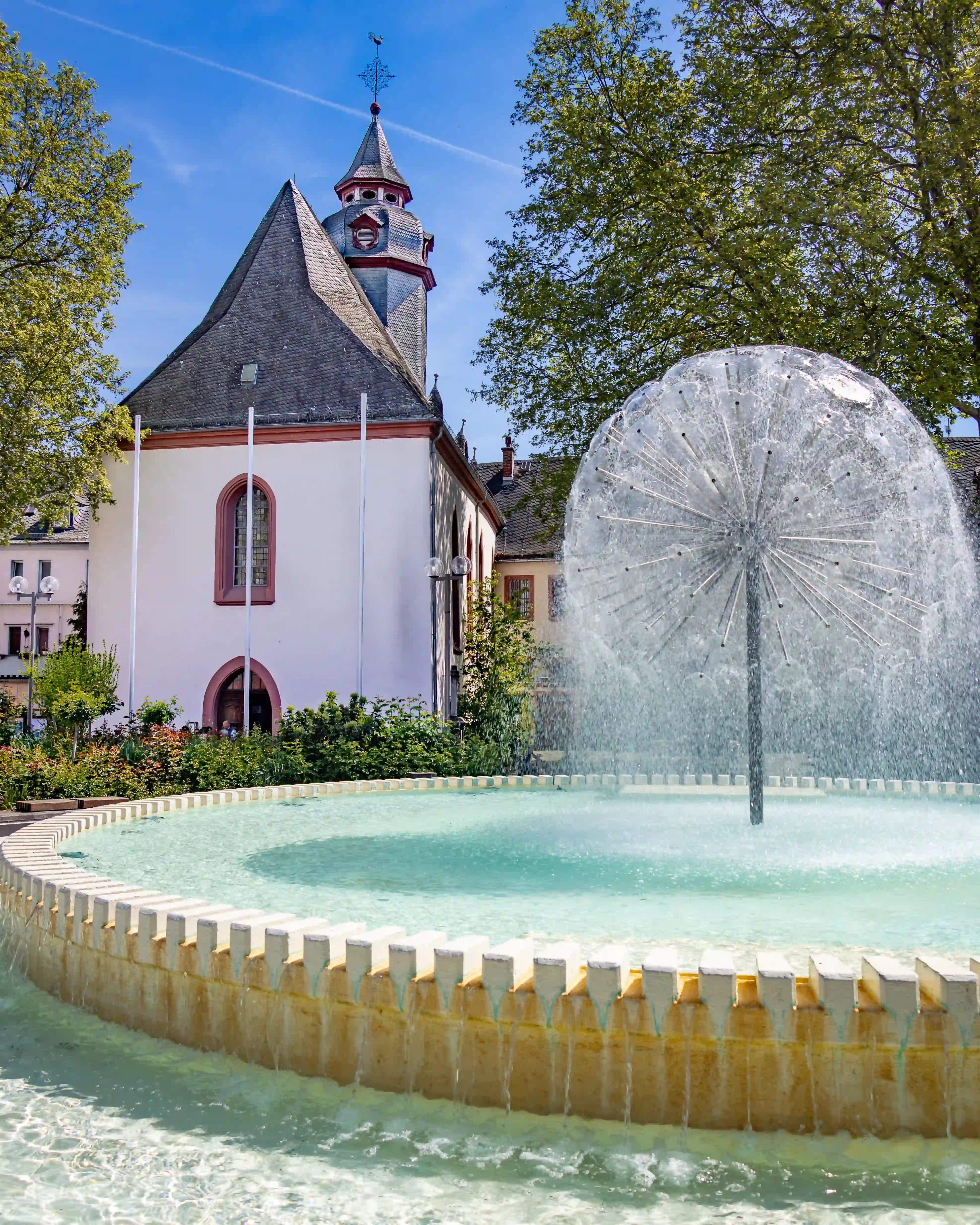 A round fountain sprays water in front of a small church and trees.