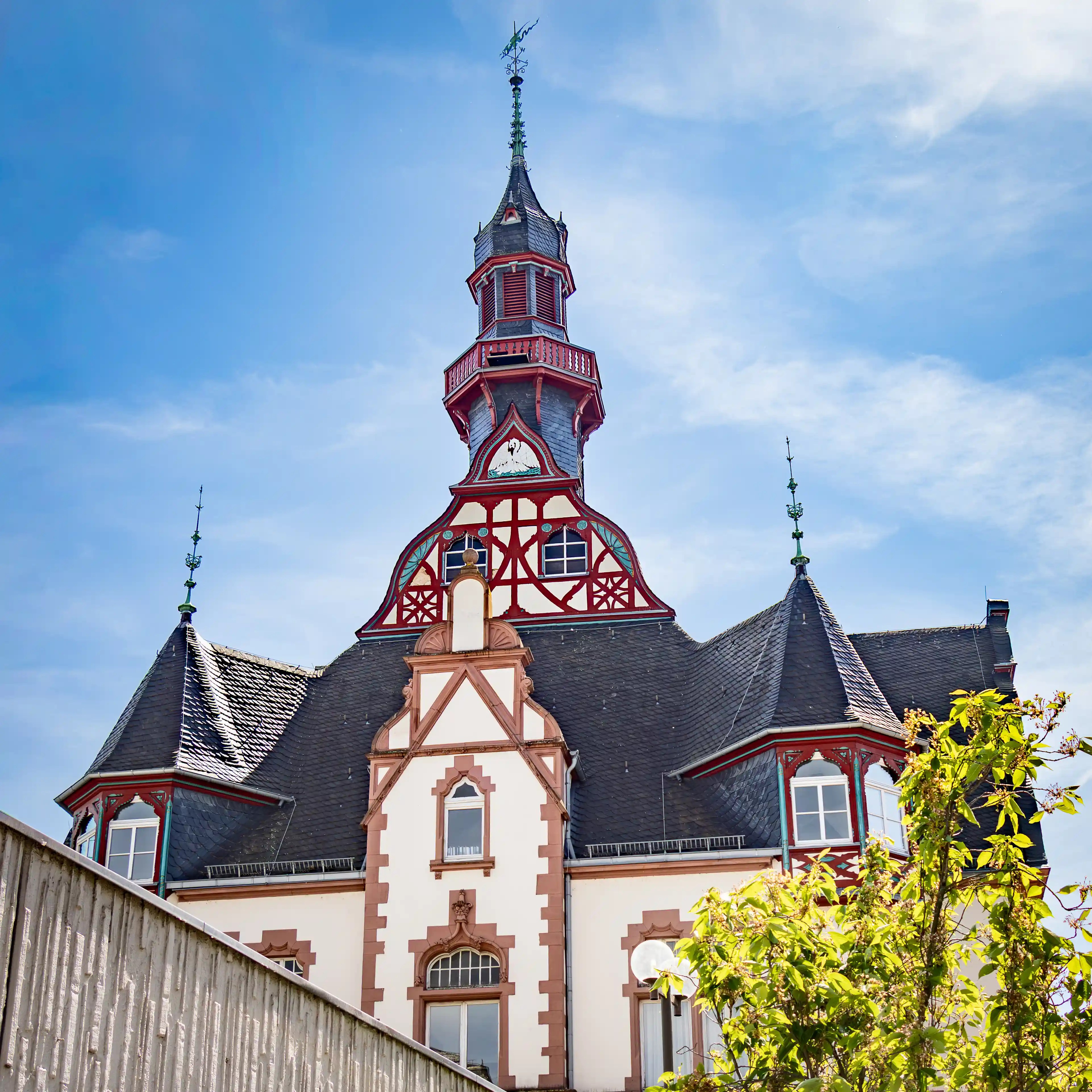 An ornate tower rises above a light-colored building with red trim and slate roofs.