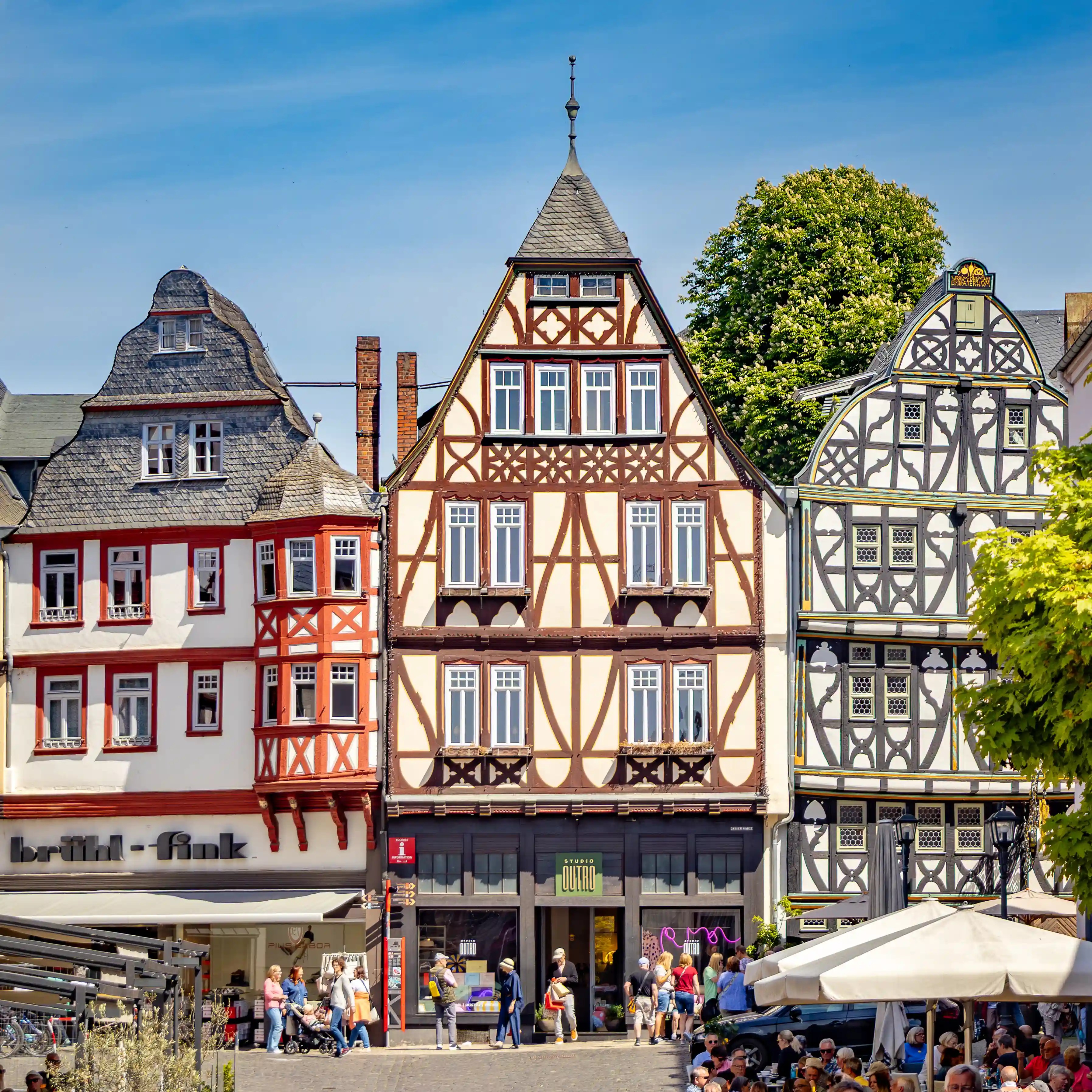 Three tall half-timbered buildings stand above a busy square with people and umbrellas.