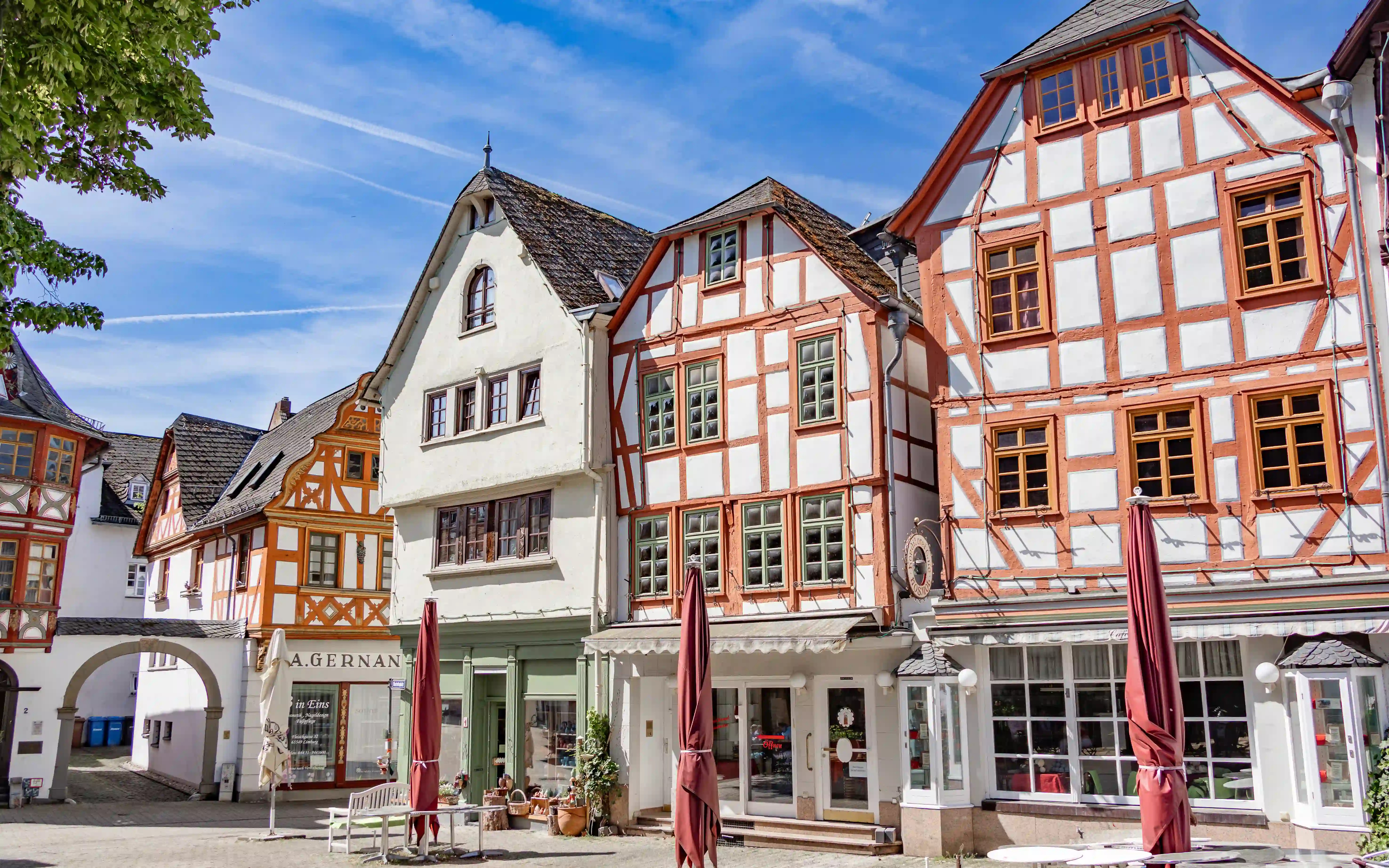 Colorful half-timbered buildings line a sunny square with closed umbrellas and shopfronts.