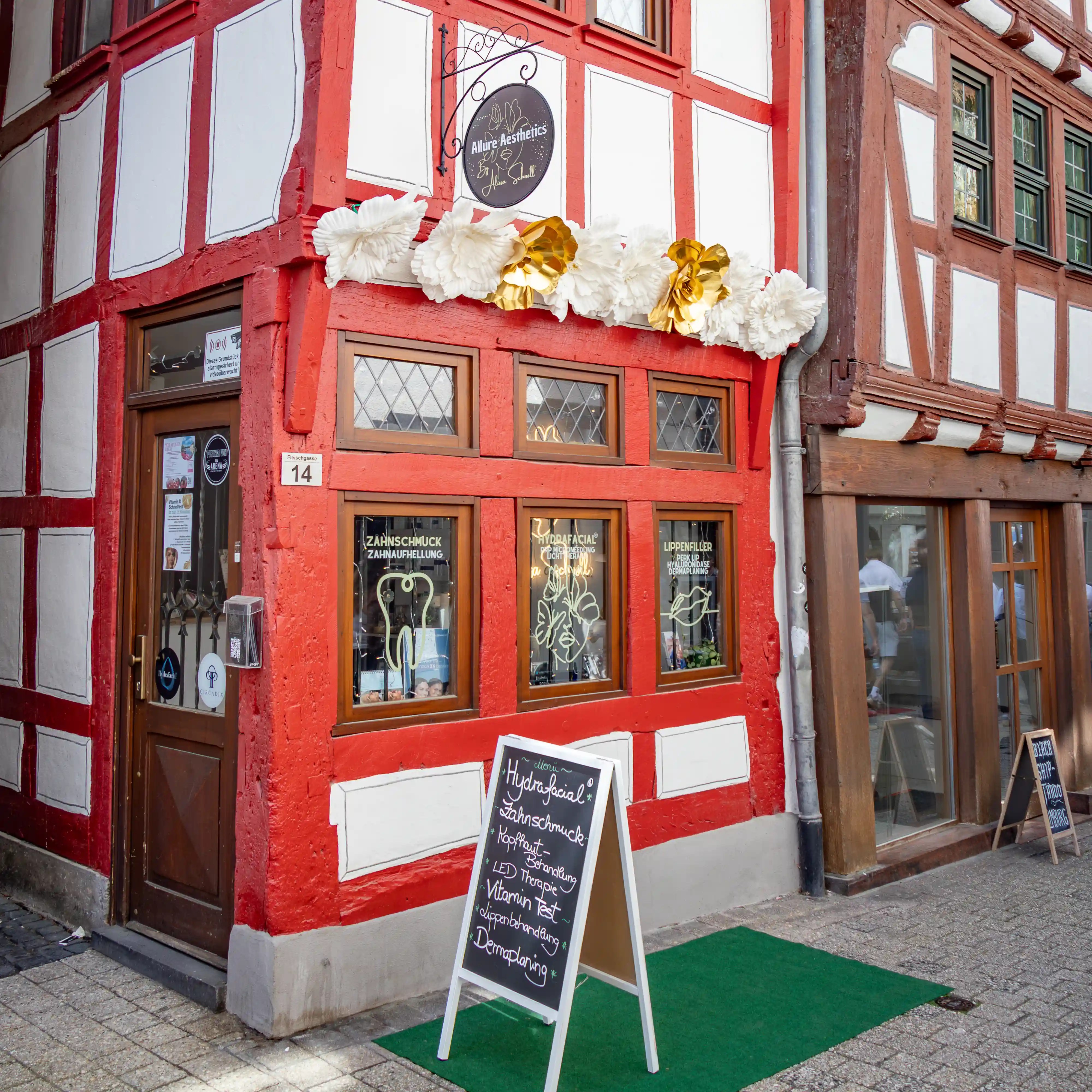 A red half-timbered storefront has large white and gold flowers above its windows.