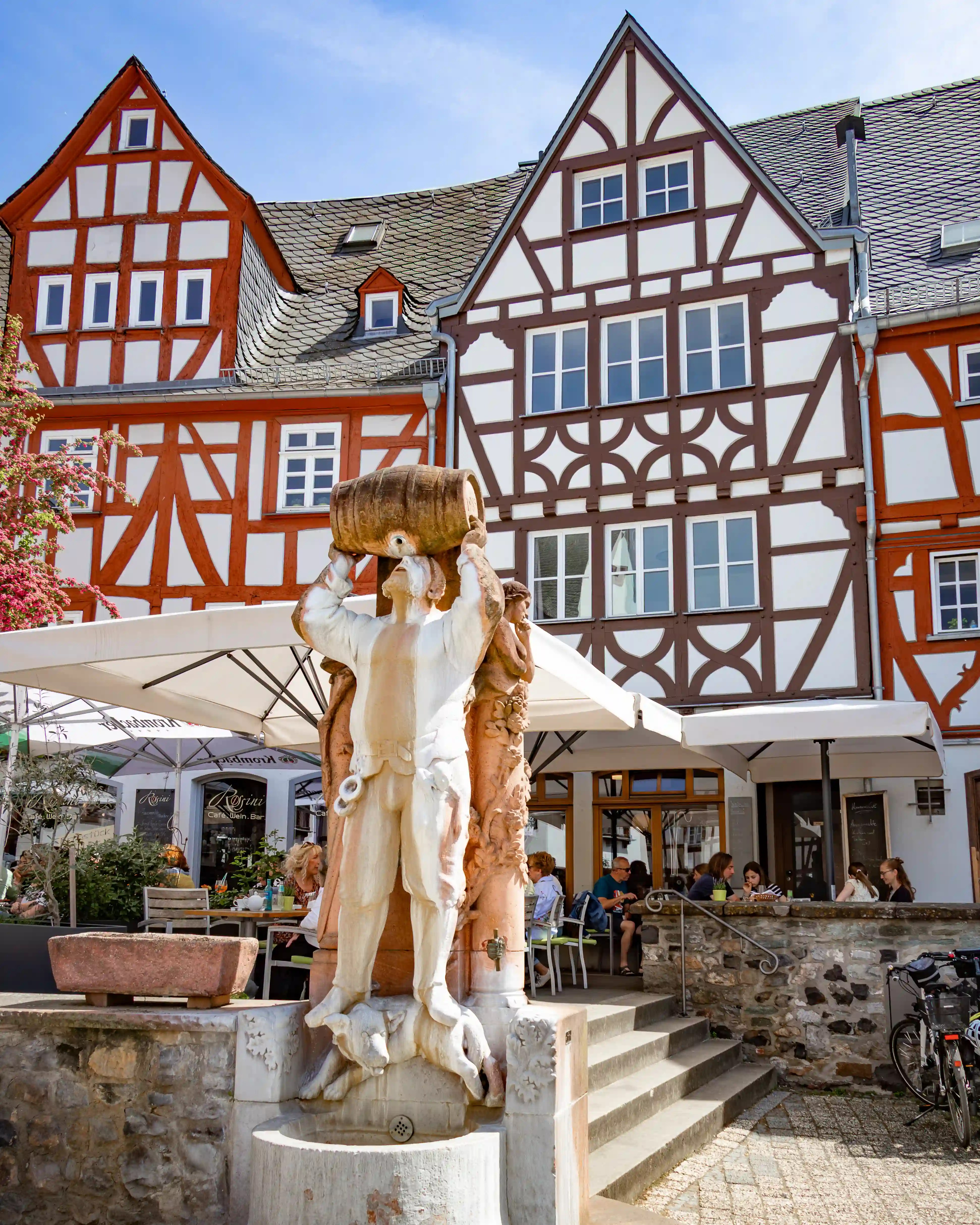 A white fountain figure holding a barrel stands in front of half-timbered buildings and cafe umbrellas.