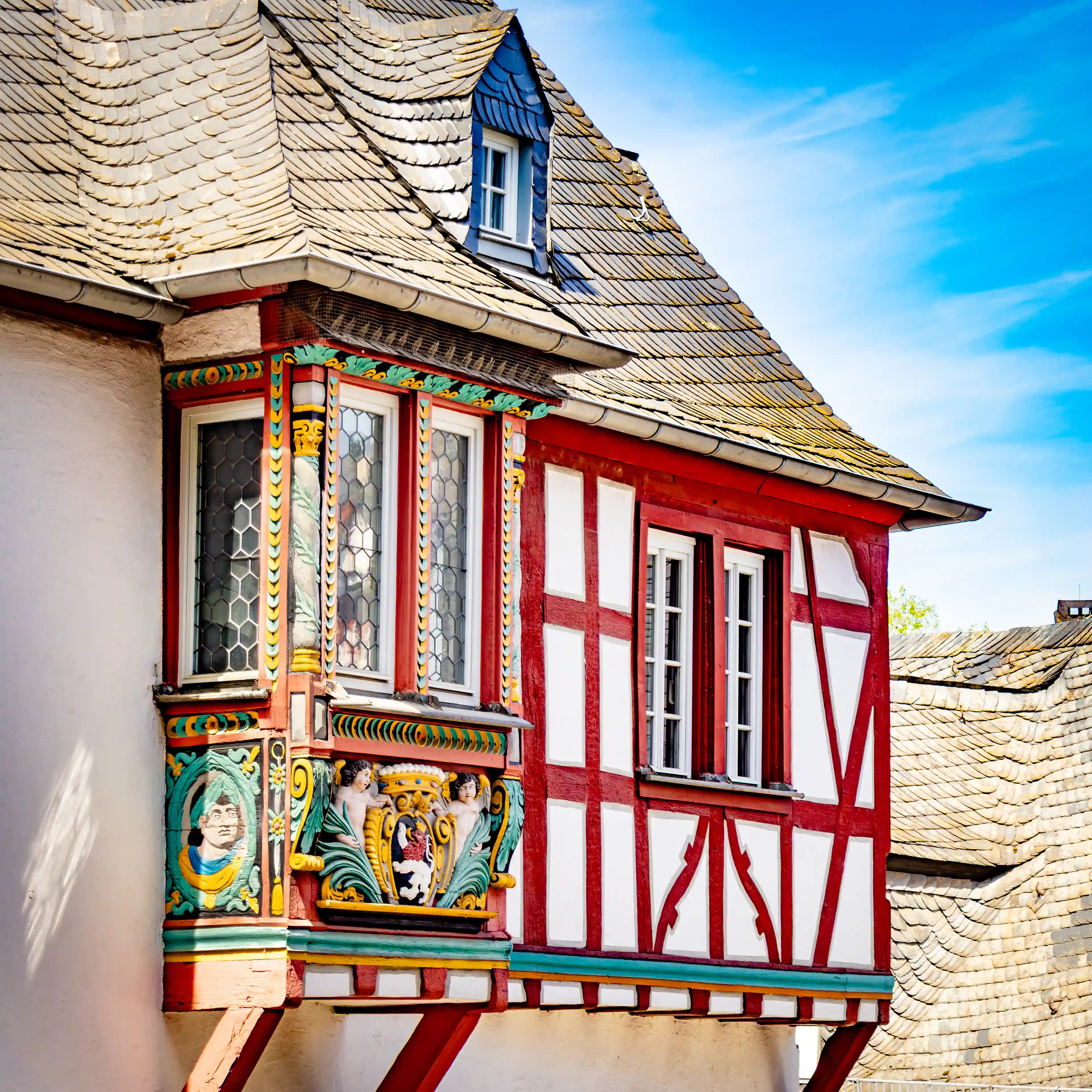 A colorful carved bay window projects from a red half-timbered building with a slate roof.
