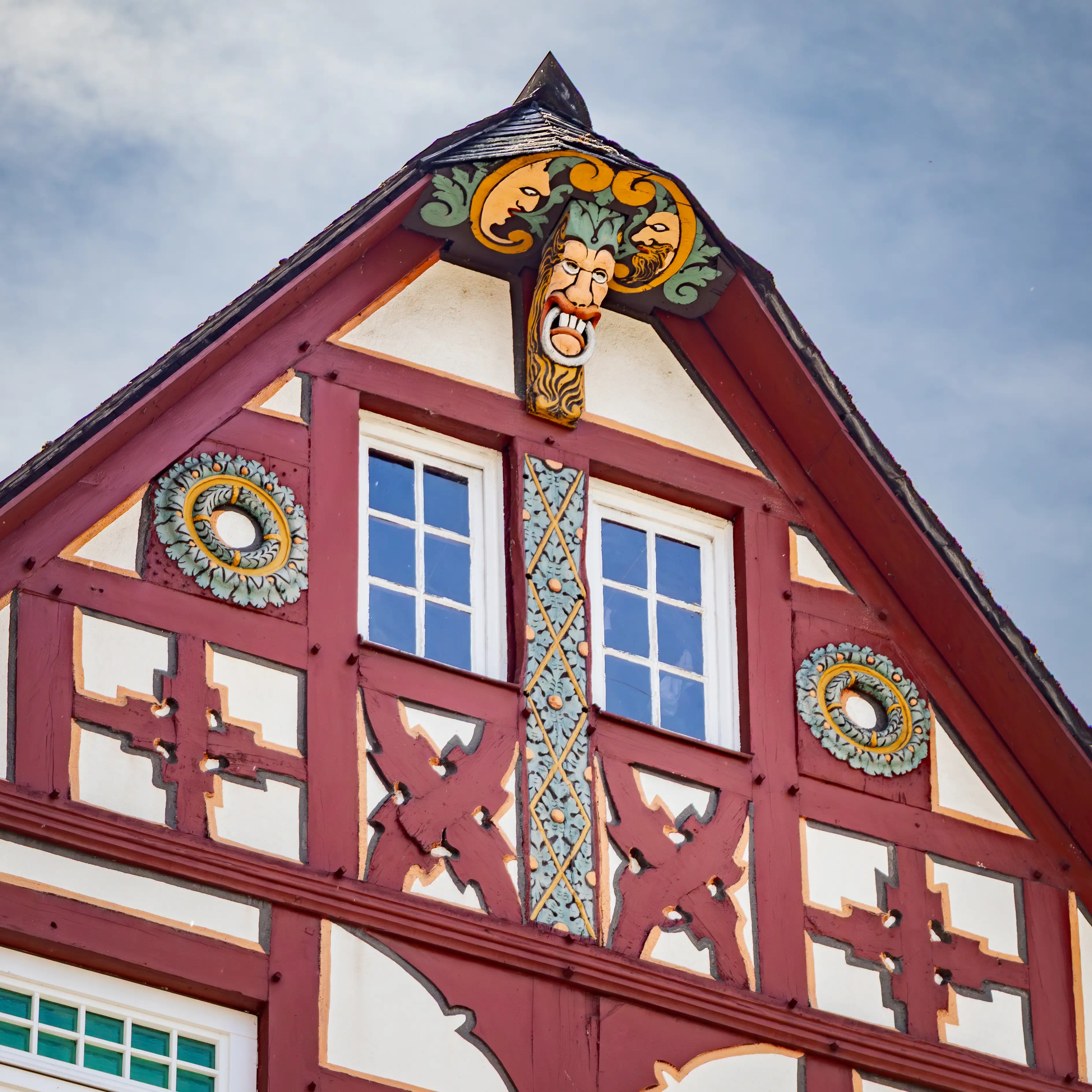 A carved face and decorative painted panels sit near the roofline of a red half-timbered building.