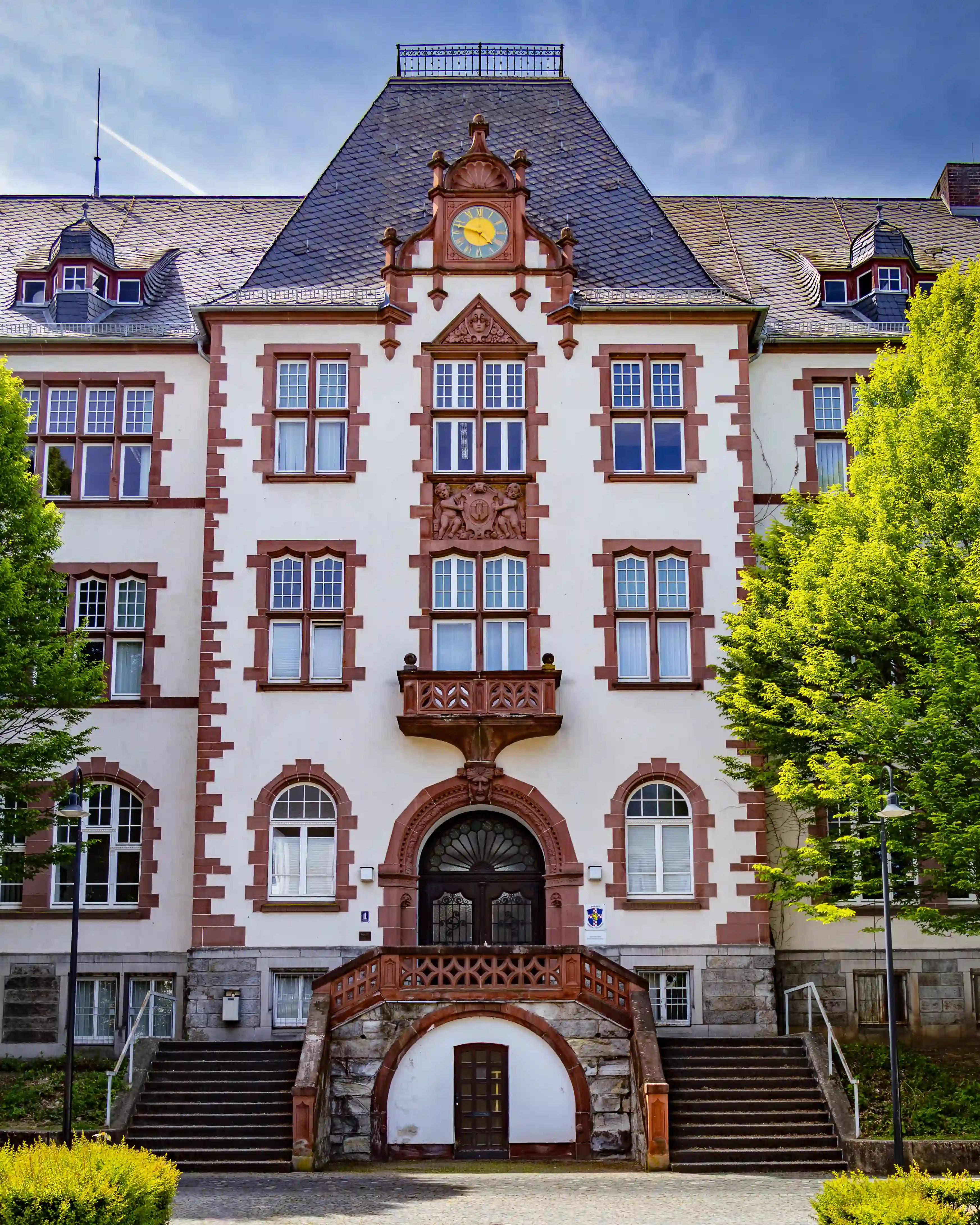 A large light-colored building has a central staircase, arched entrance, balcony, and clock.