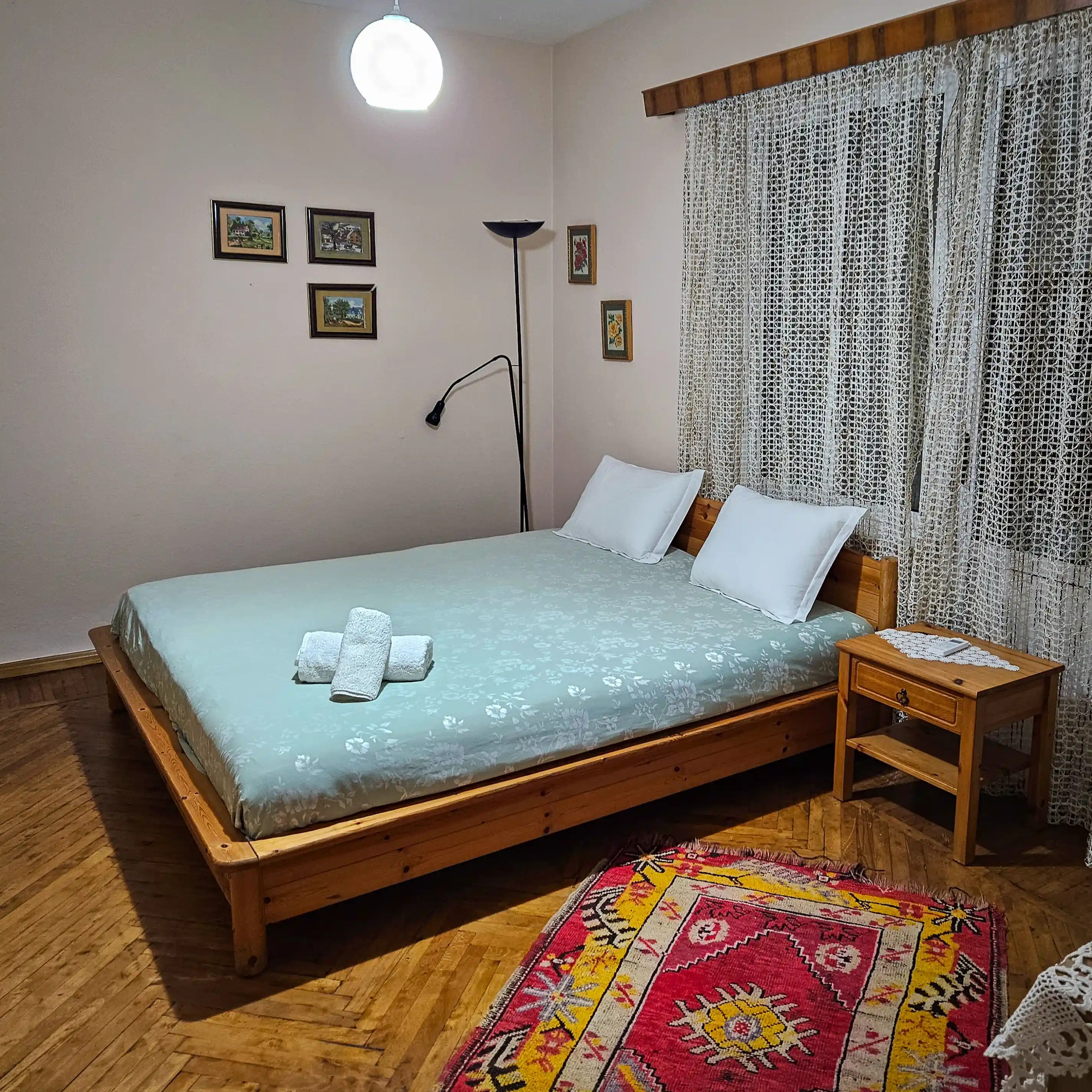Bedroom with a wooden bed, lace curtains, and a colorful rug in an apartment in Berat.