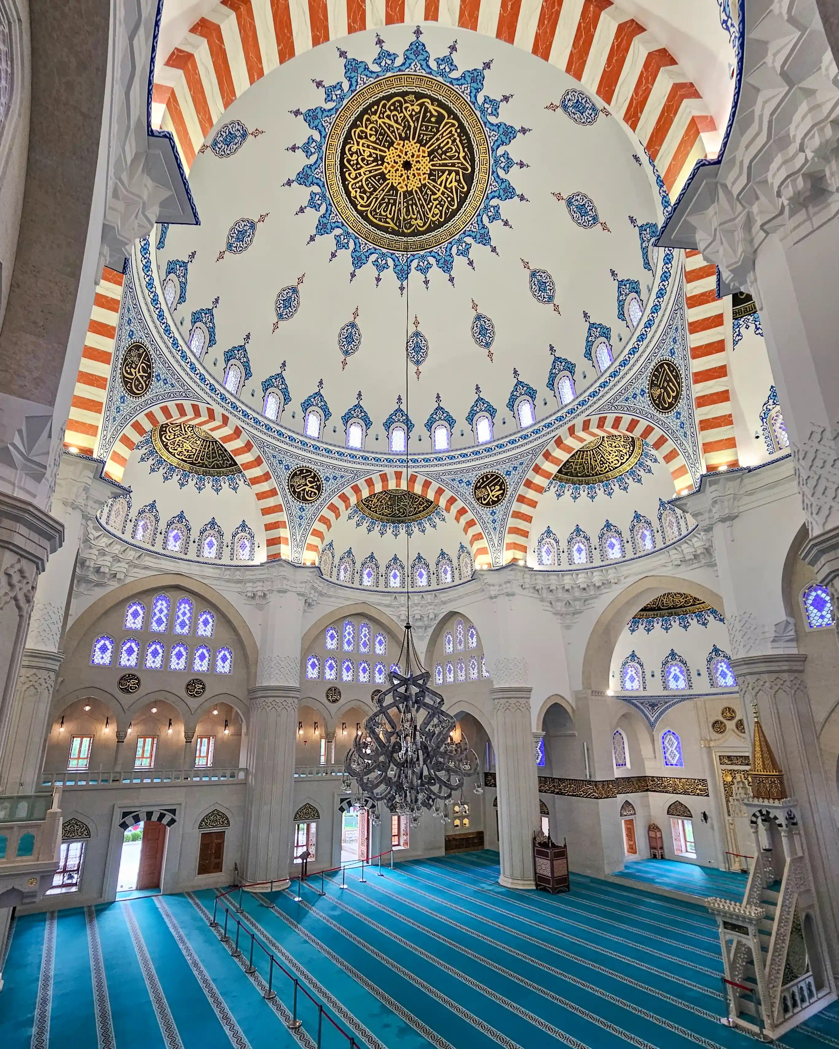 Interior of the Grand Mosque of Tirana with a large central dome, chandelier, and blue prayer carpet.