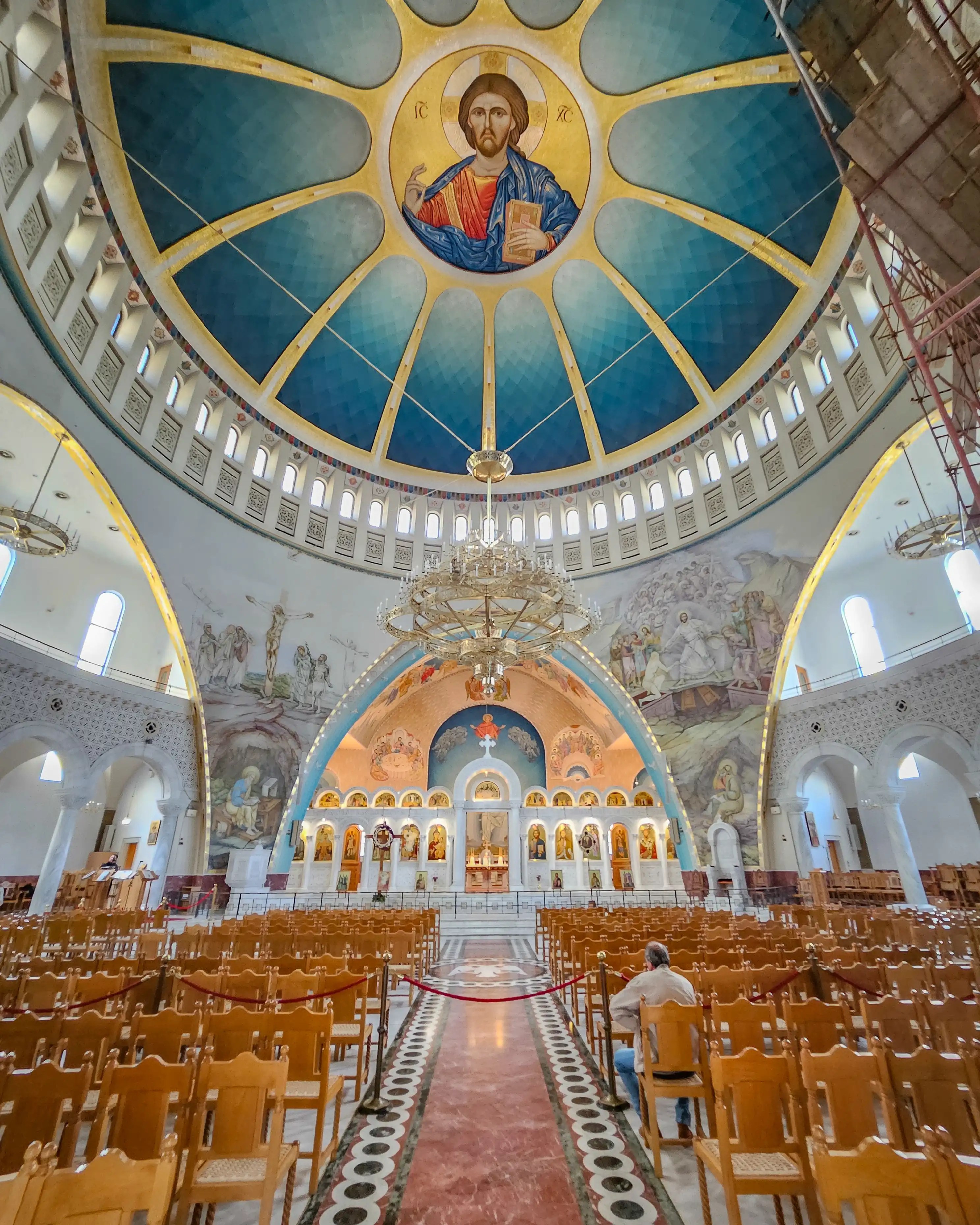 Interior of the Resurrection of Christ Orthodox Cathedral in Tirana with a large blue dome, icon of Christ, and rows of wooden chairs.