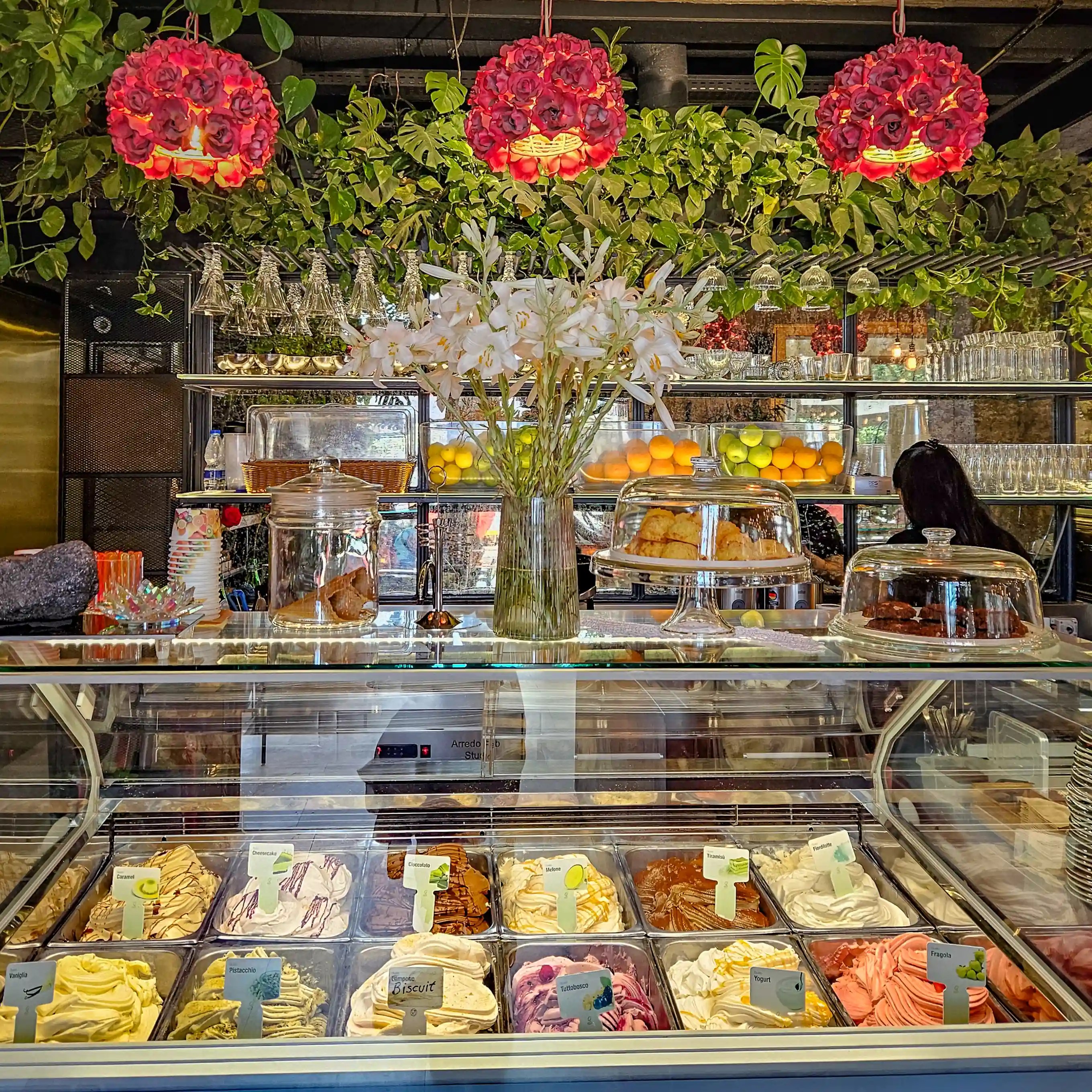 A display case filled with gelato beneath flowers, plants, and shelves of fruit inside Café Botanica in Tirana.
