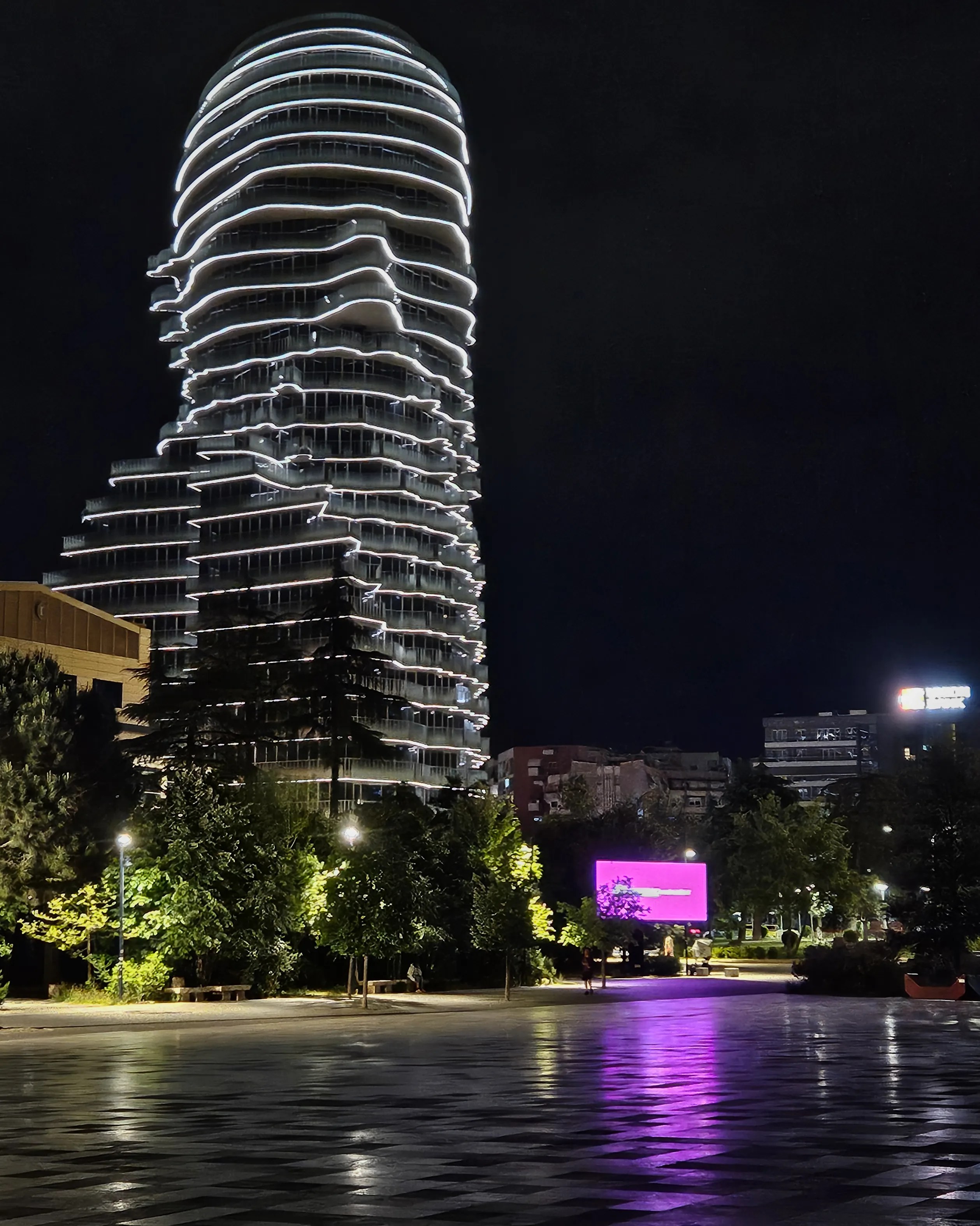 A modern high-rise tower in the form of a face outlined with white lights rises above a park at night in Tirana.