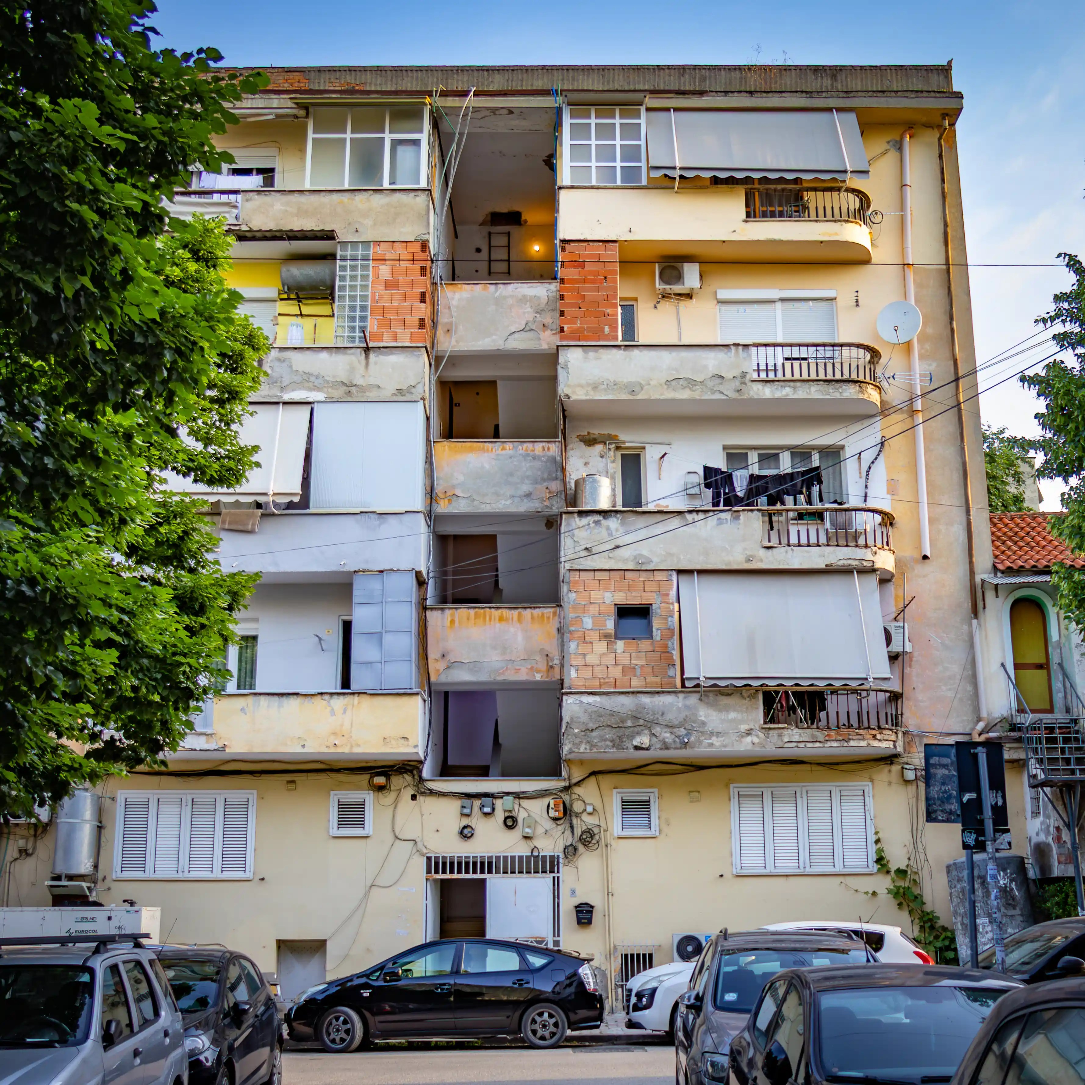 A mid-rise apartment building in Tirana with balconies, weathered exterior walls, and parked cars along the street.
