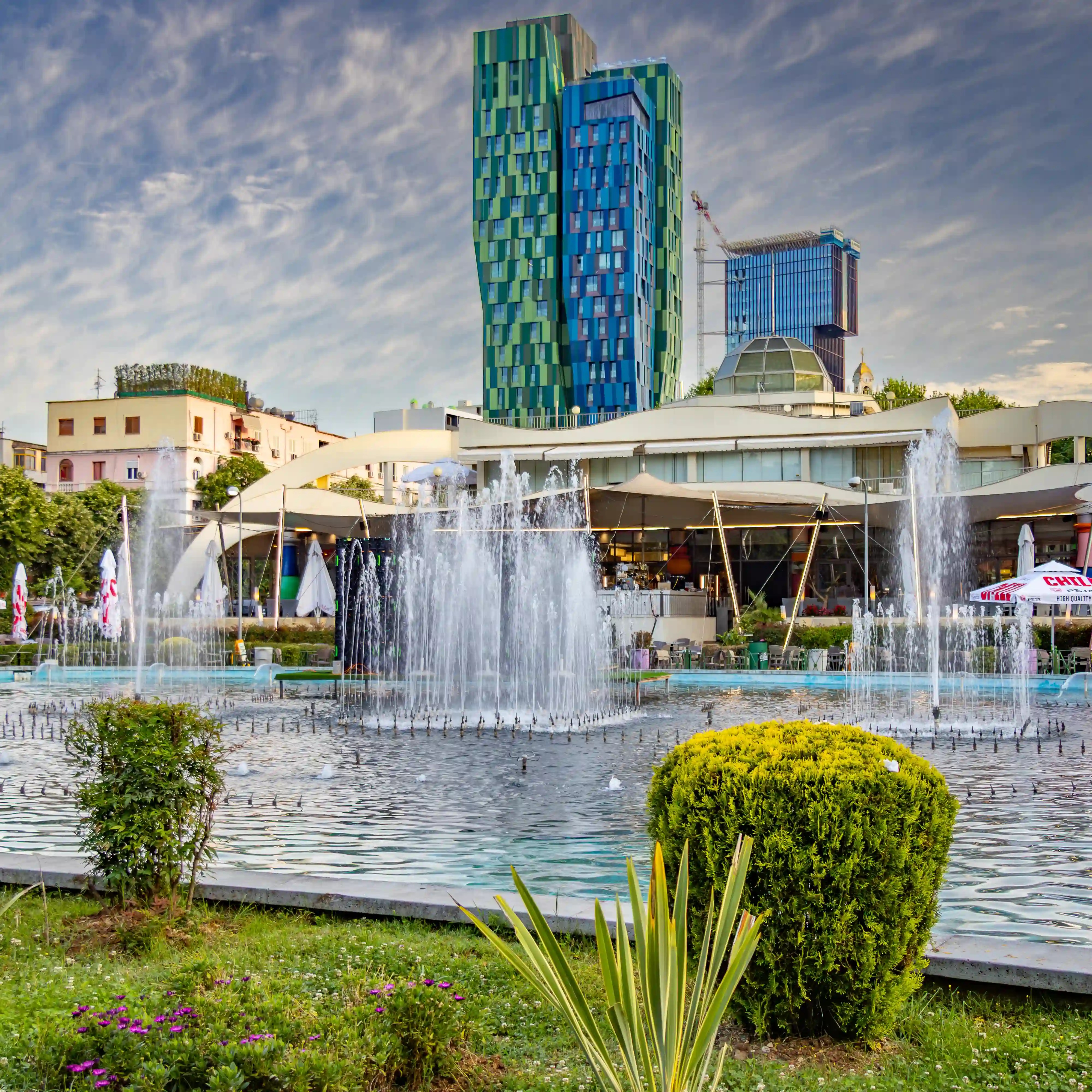 Fountains spray into a large pool with colorful modern towers rising in the background in central Tirana.
