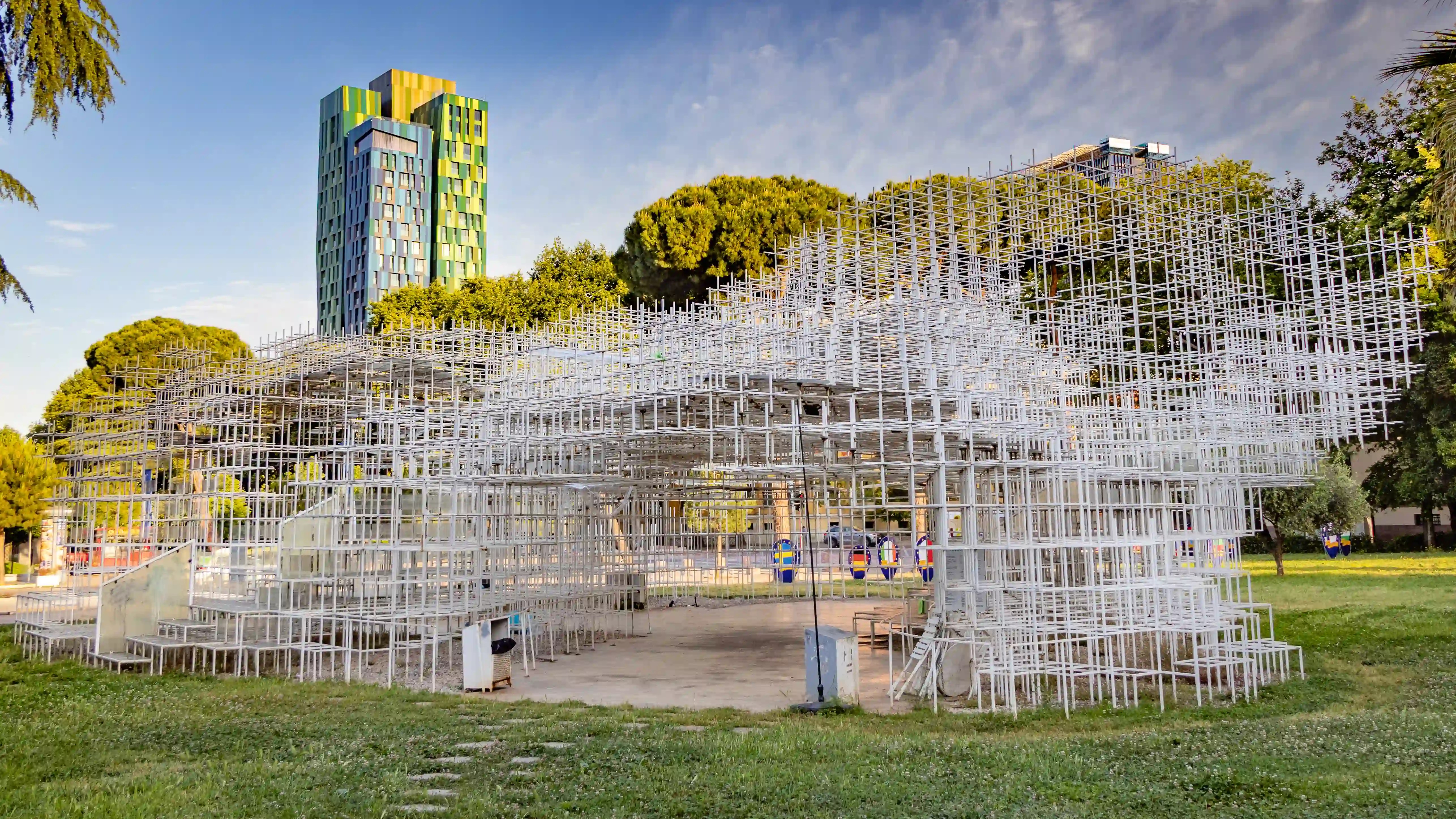 The Cloud installation in Tirana, an open steel lattice structure standing in a grassy urban park with a colorful modern tower in the background.