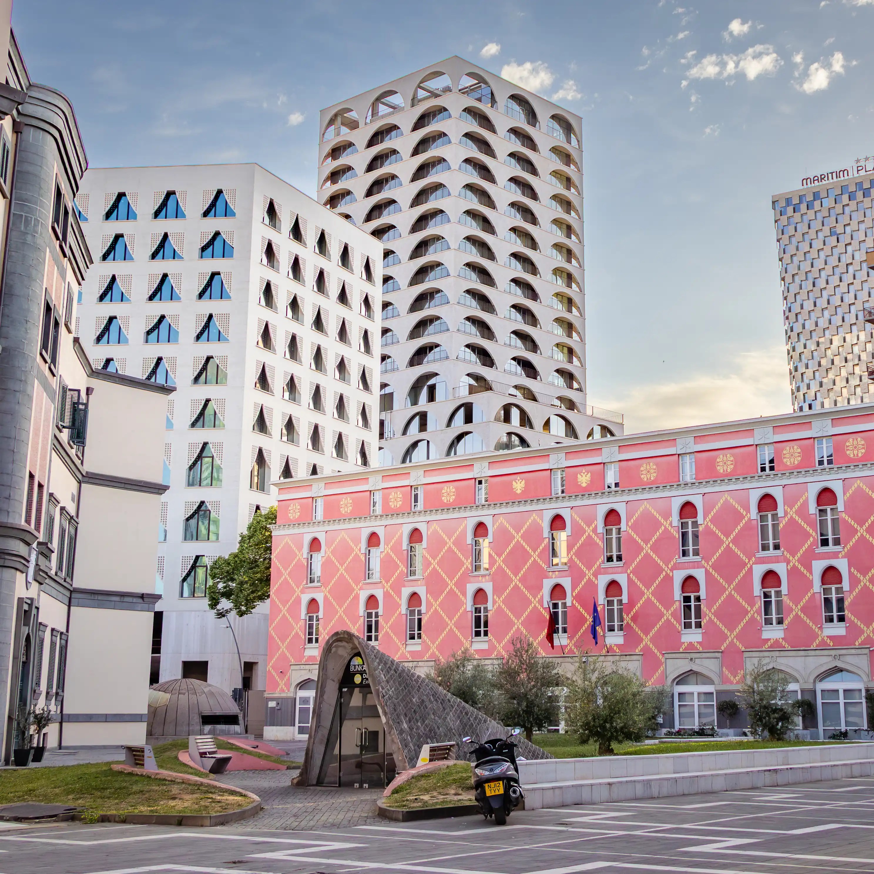 A pink historic building stands beside modern white high-rise towers in central Tirana.