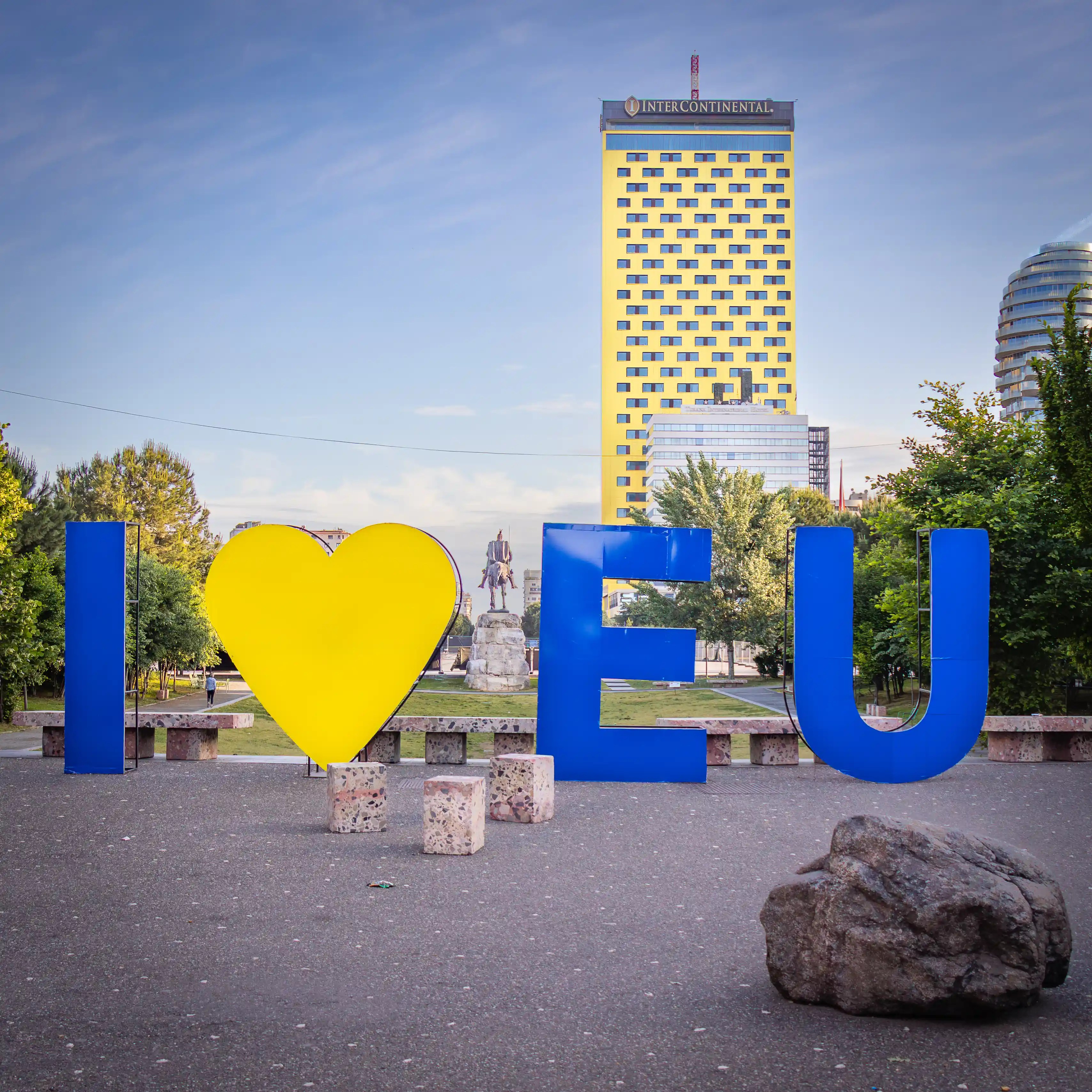 A large blue and yellow "I ♥ EU" sign stands in a city square with buildings and a statue visible behind it.