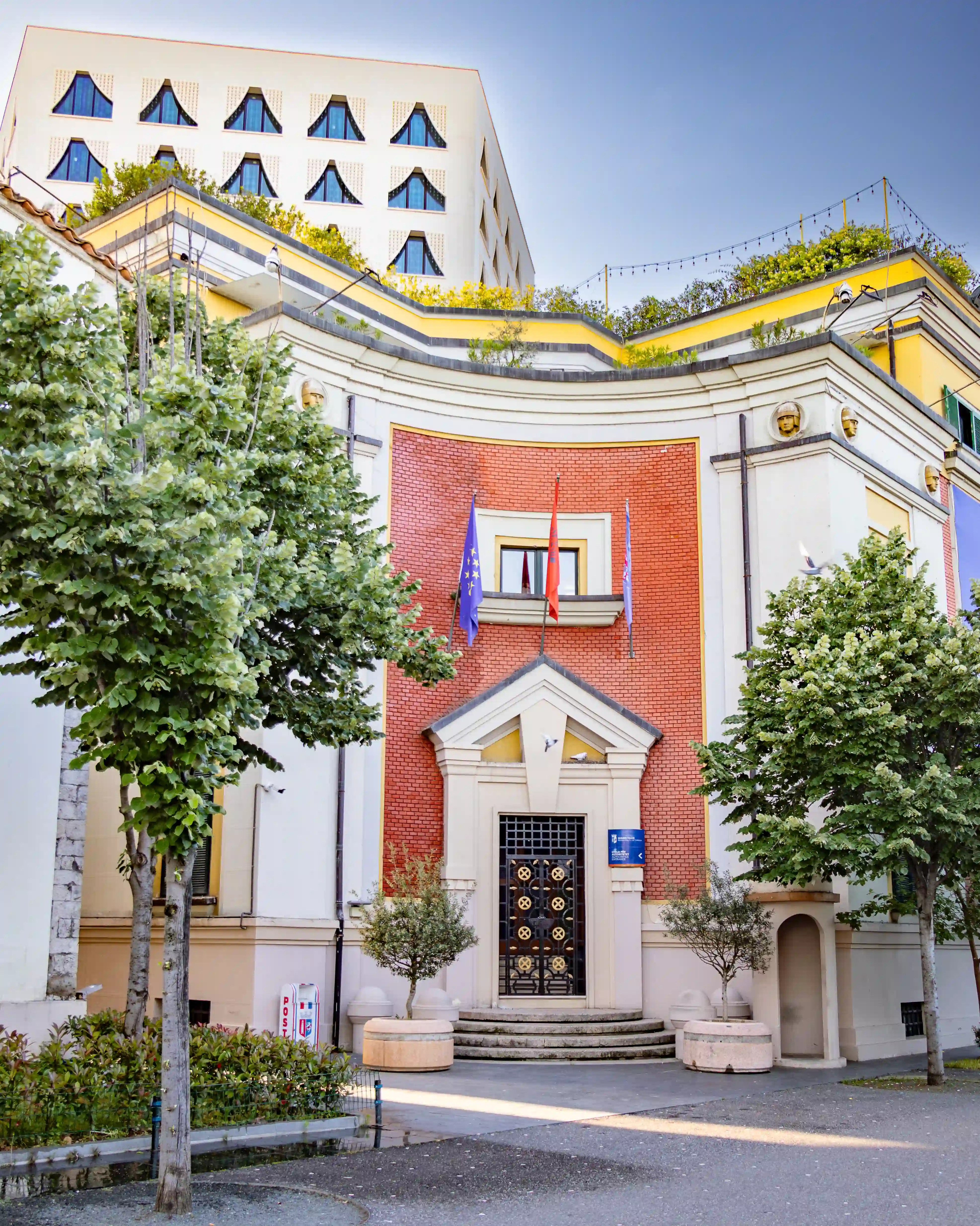 A historic building displaying Albanian and European Union flags stands beneath a modern white building in central Tirana.