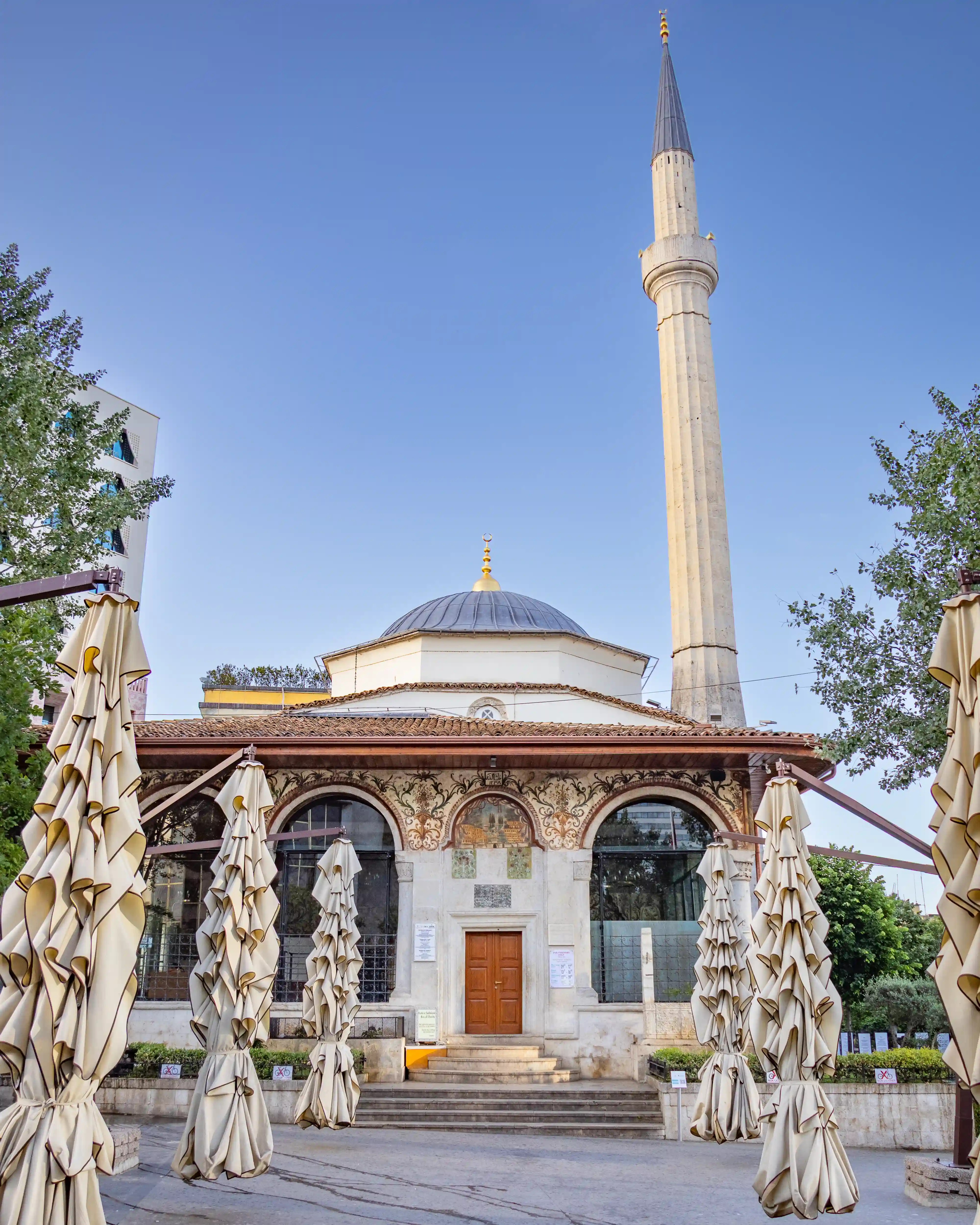 Front entrance of the Et'hem Bej Mosque with its dome and minaret rising above closed shade umbrellas.