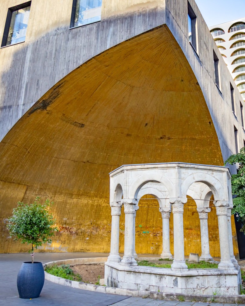 A small white stone pavilion with arched columns stands in front of a massive modern building with a curved golden concrete wall in central Tirana.