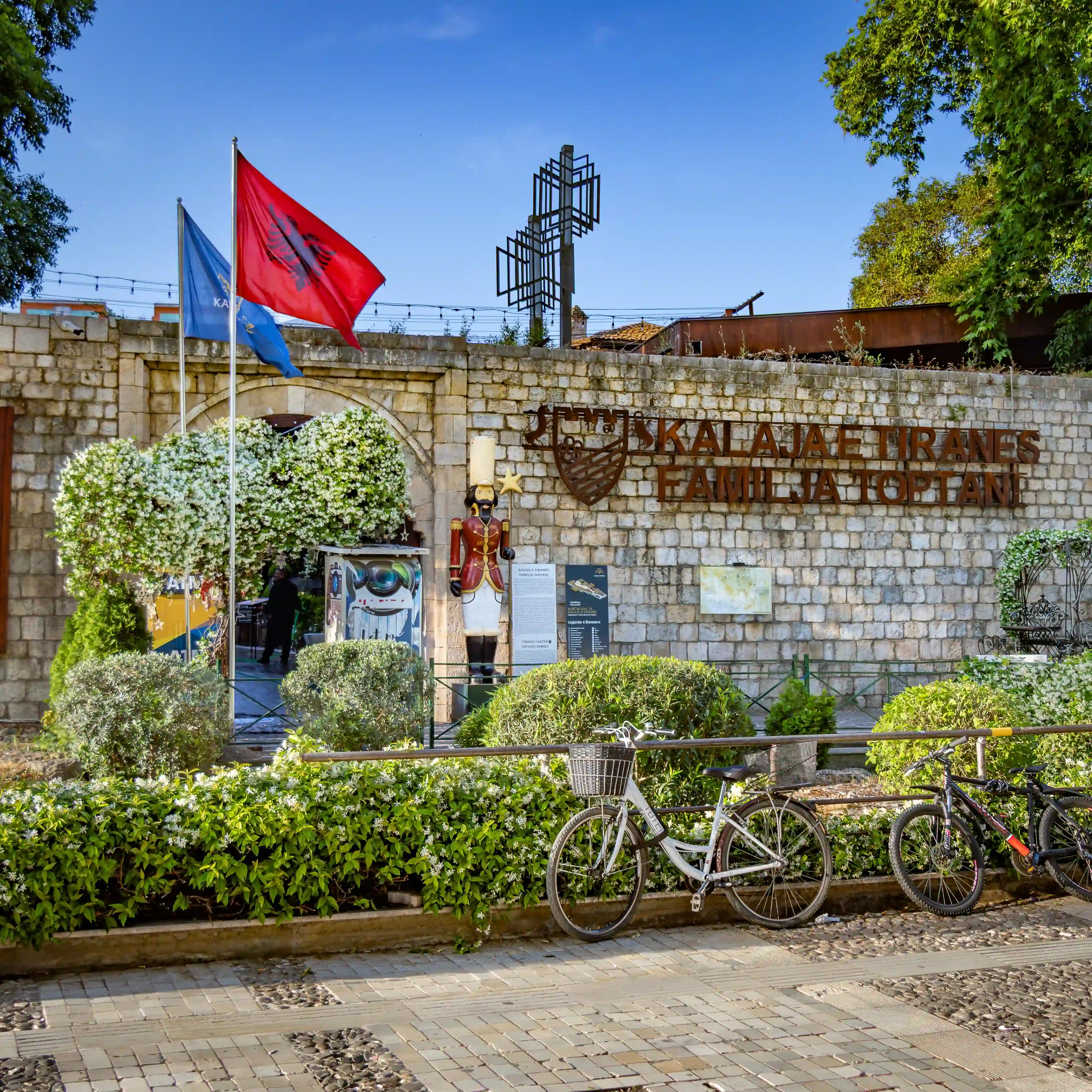 The entrance to Tirana Castle with Albanian flags, stone walls, and the castle sign above a historic gateway.