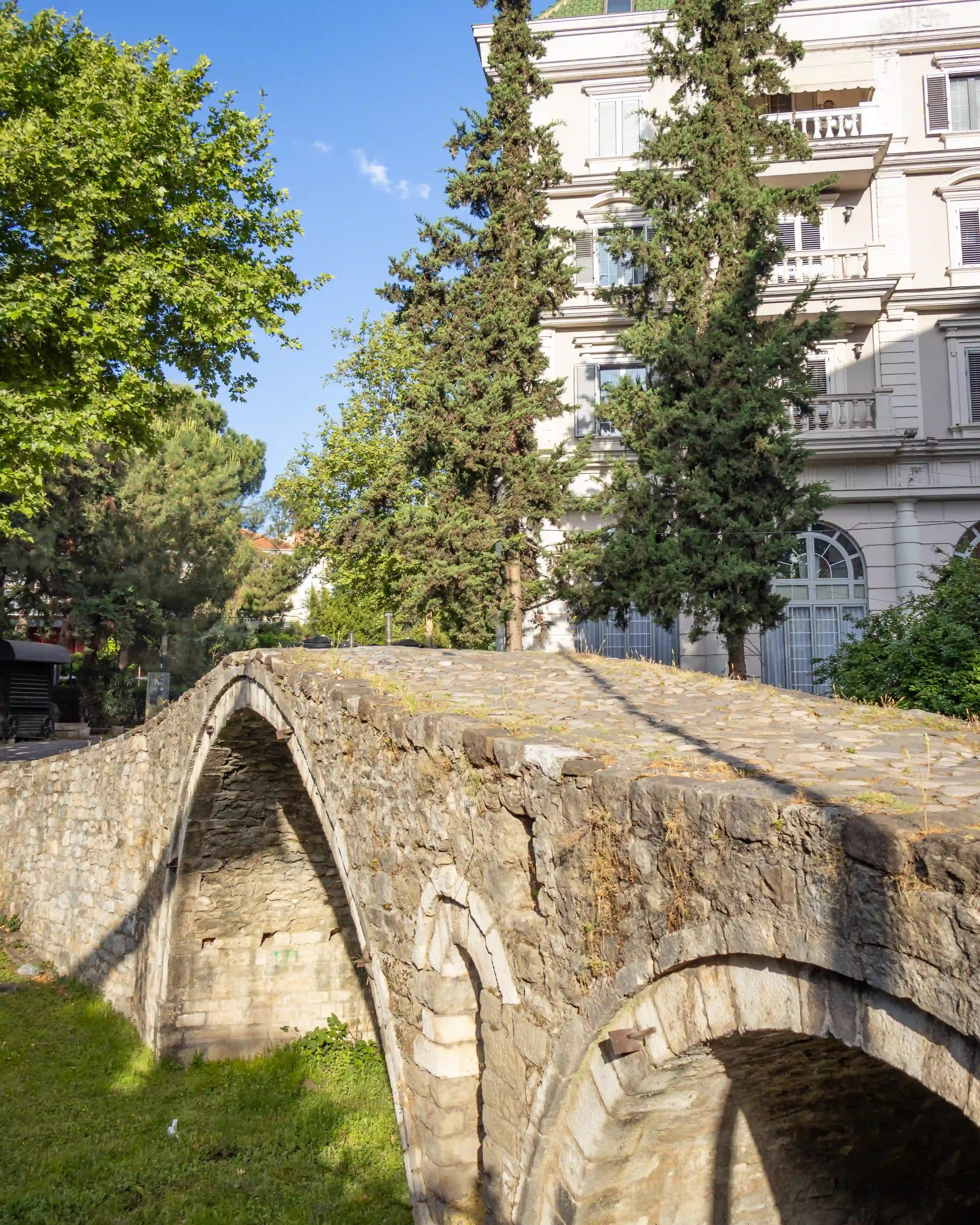 Side view of the stone Tanners' Bridge in Tirana with nearby historic villas and trees.